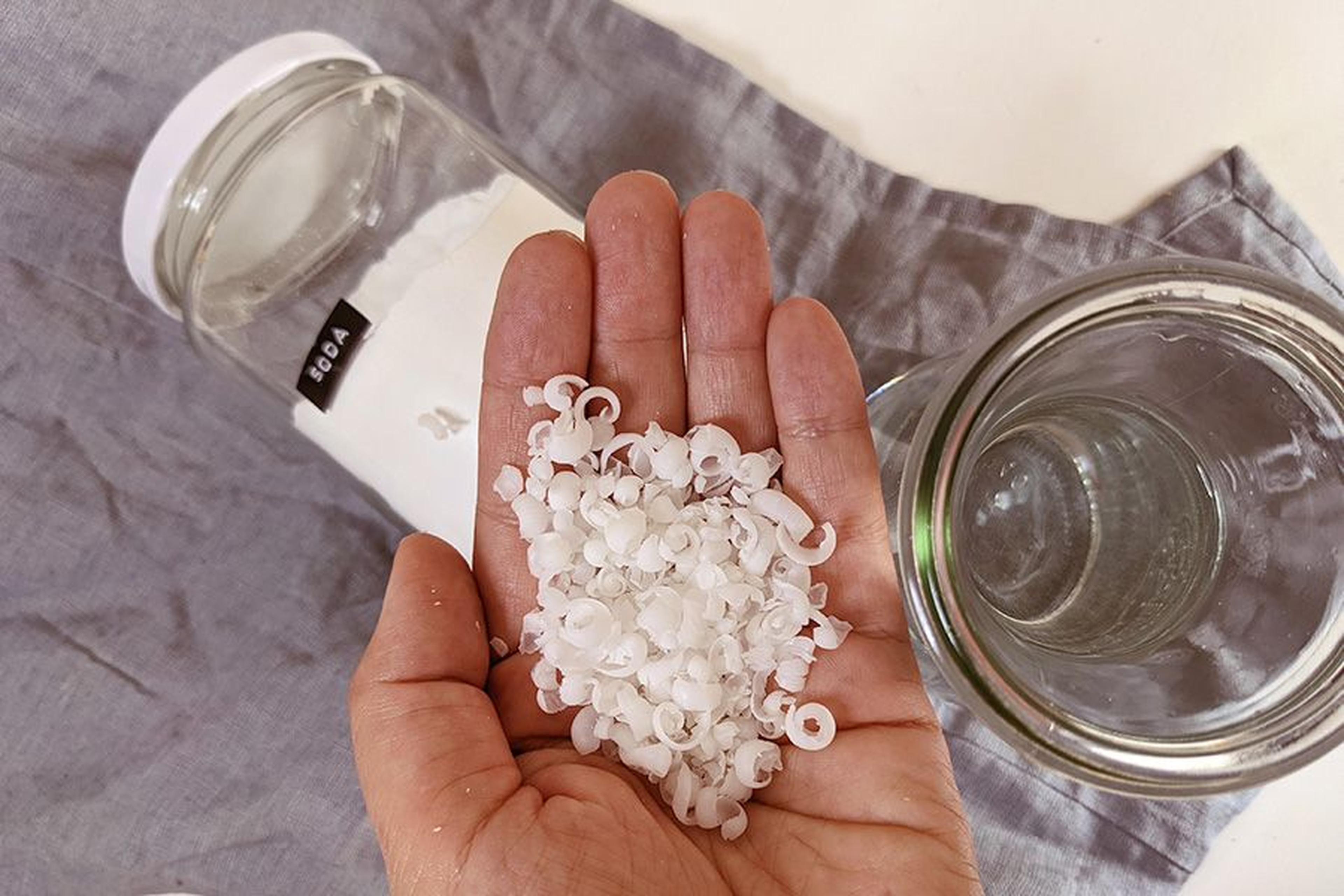 Hand, die kleine weiße Seifenraspeln über einem grauen Tuch hält, daneben ein Sodaglas und ein leerer Behälter.