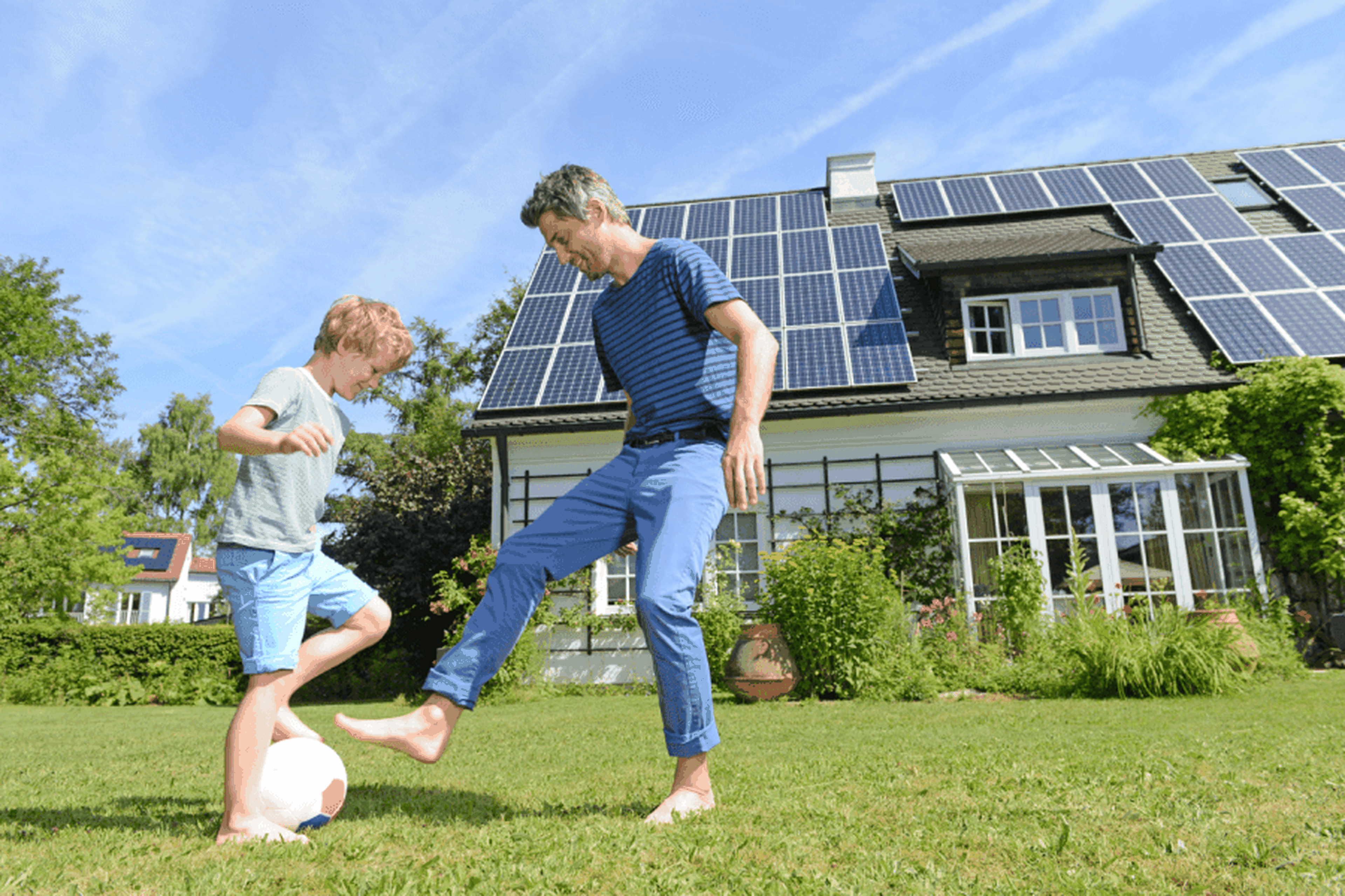 Vater und Sohn spielen barfuß Fußball auf dem Rasen vor einem Haus mit Sonnenkollektoren und einem Garten.