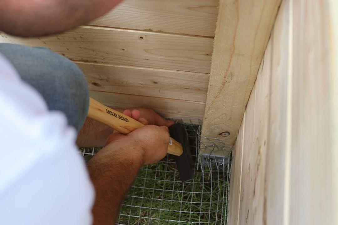 Person, die mit einem Hammer Drahtgeflecht an der Unterseite von Holzplatten in einem umzäunten Bereich im Freien befestigt.