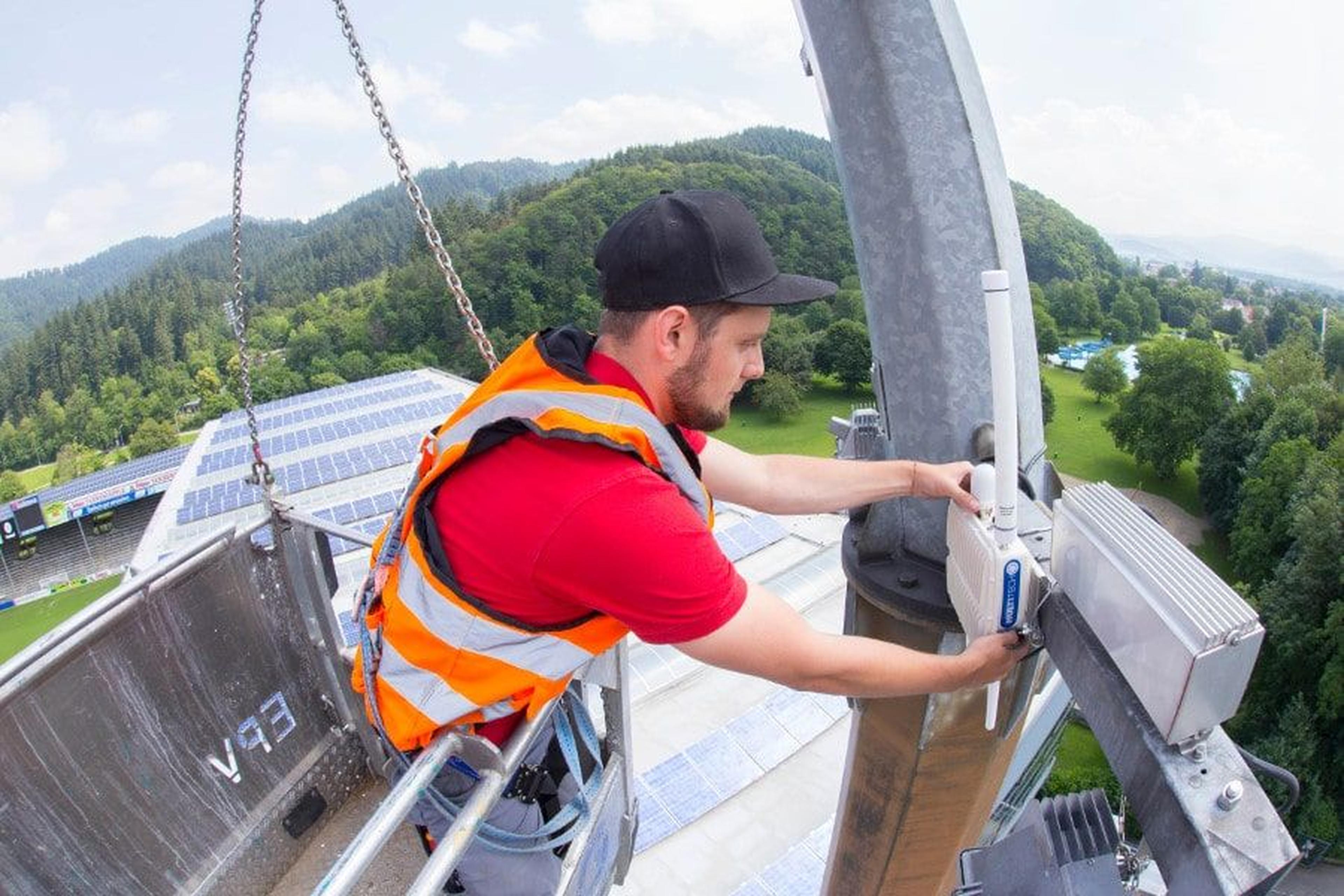 Ein Arbeiter in Sicherheitsweste und Schutzhelm installiert Geräte auf einem hohen Bauwerk mit Blick auf ein Solarpanel-Feld und bewaldete Hügel.