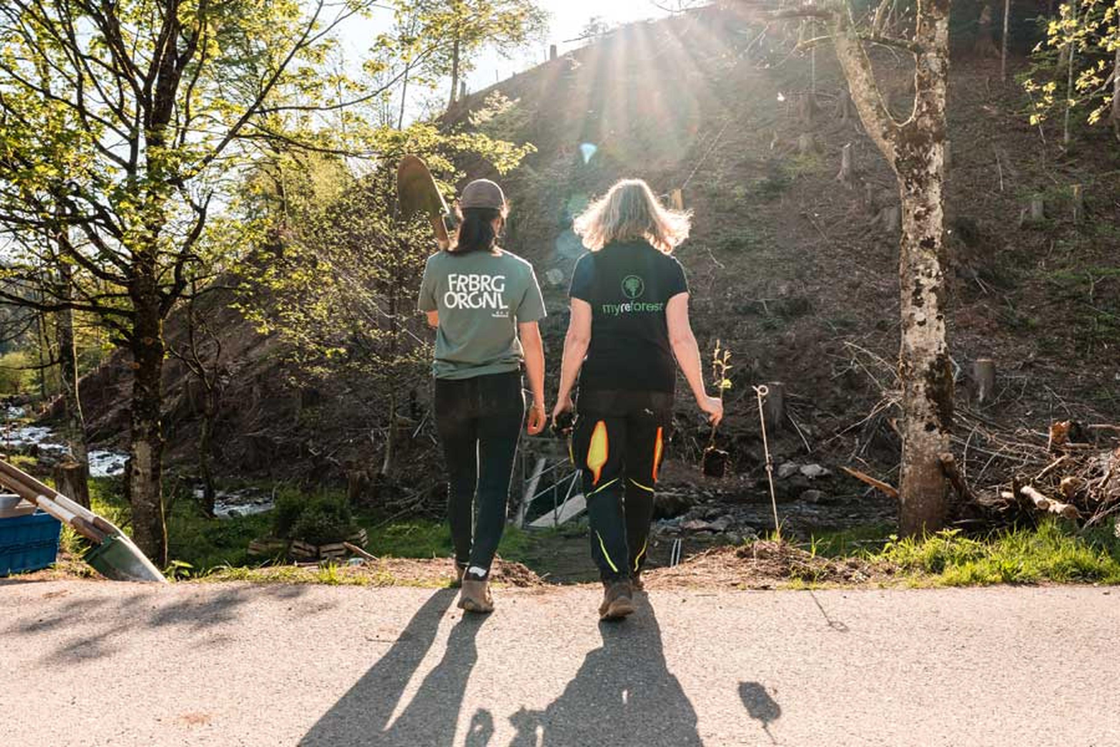Zwei Personen gehen auf einem Weg, sie halten Schaufeln und kleine Pflanzen in den Händen, während Sonnenlicht durch die Bäume in einer natürlichen Umgebung scheint.