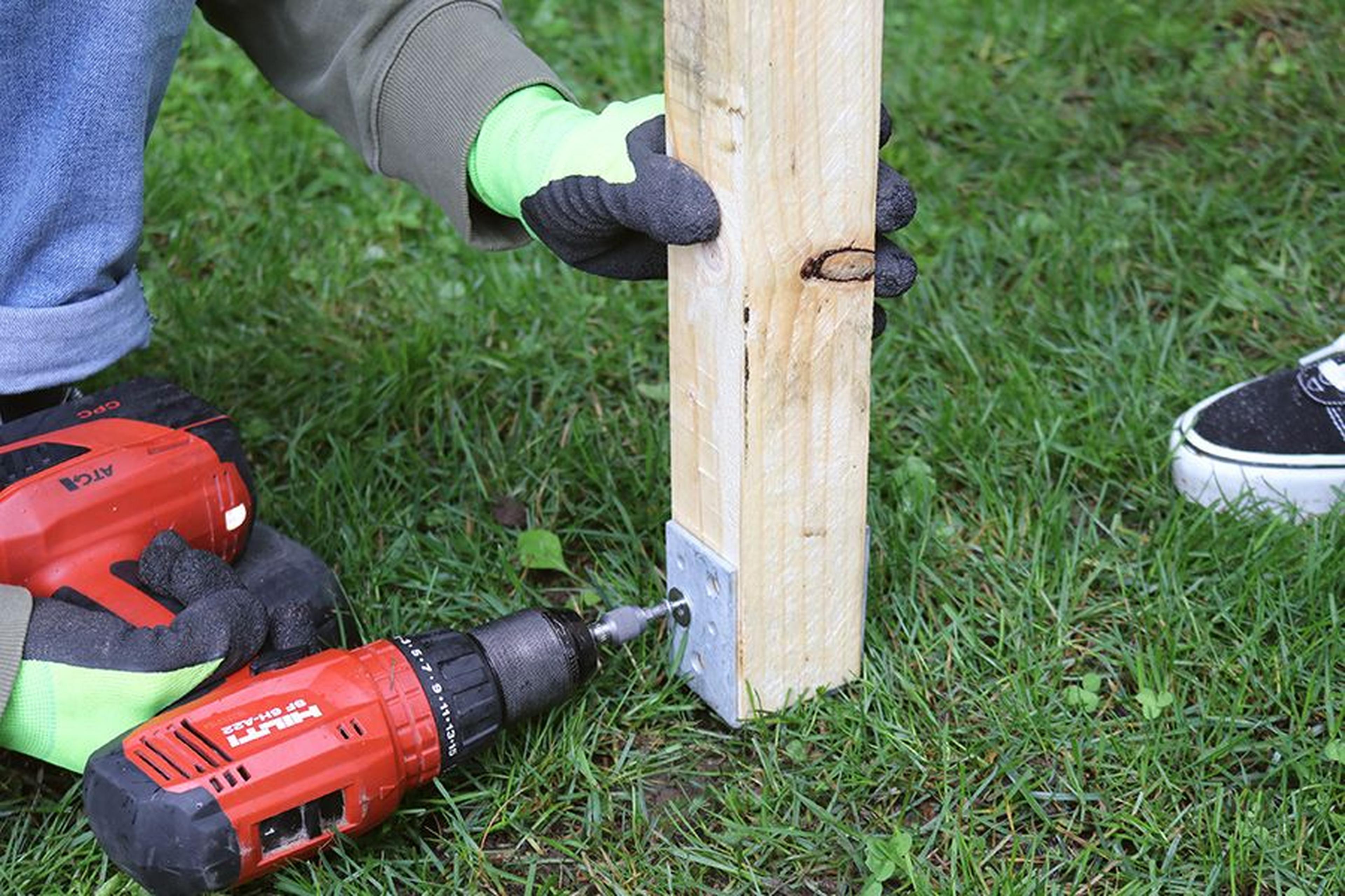 Person, die mit einer roten Handbohrmaschine einen Holzpfosten mit einer Metallhalterung auf Gras befestigt und dabei grüne Handschuhe trägt.