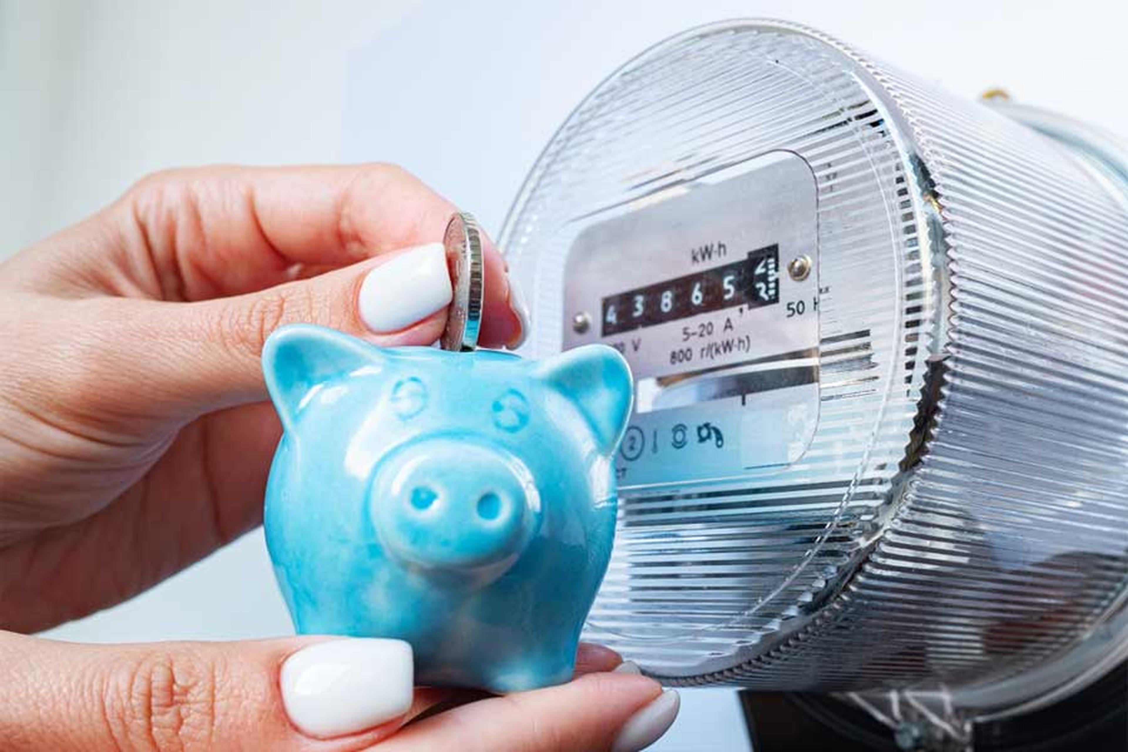 Eine Frauenhand steckt eine Geldmünze in ein kleines blaues Sparschwein, ein Stromzähler im Hintergrund