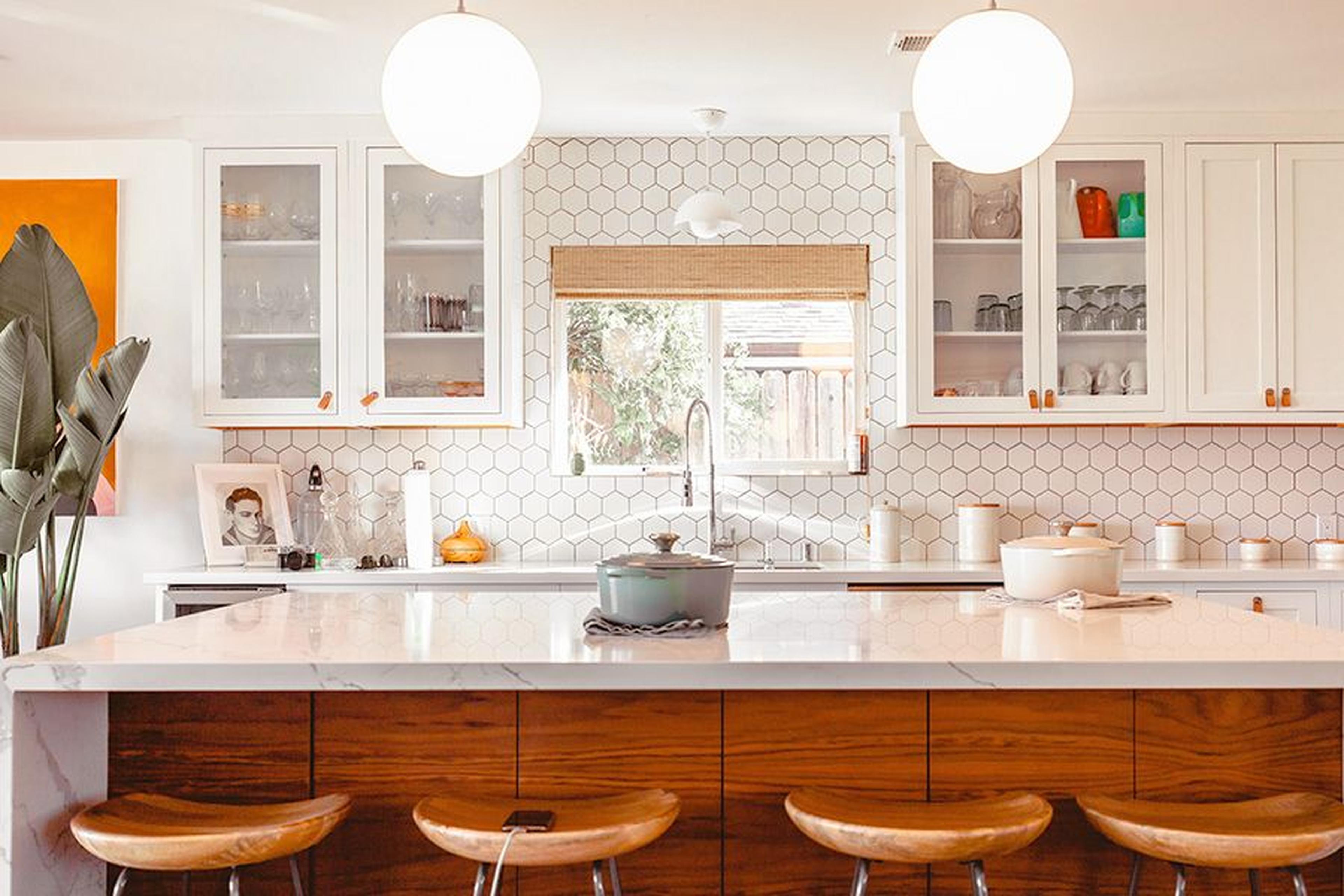 Moderne Küche mit sechseckigen Fliesen als Spritzschutz, Holzinsel, weißen Schränken und kugelförmigen Pendelleuchten. Ein Fenster bietet Blick auf den Garten.