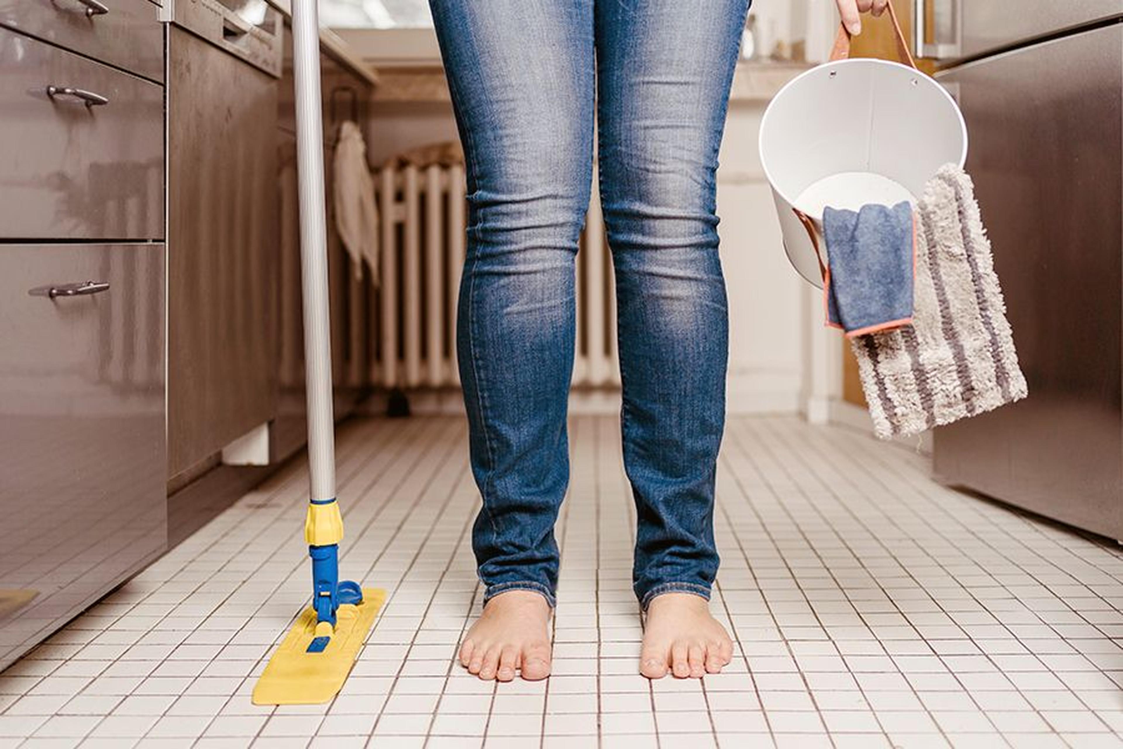 Person, die barfuß auf einem gefliesten Küchenboden steht und einen Moppeimer und einen Mopp in der Hand hält, bereit zum Putzen.