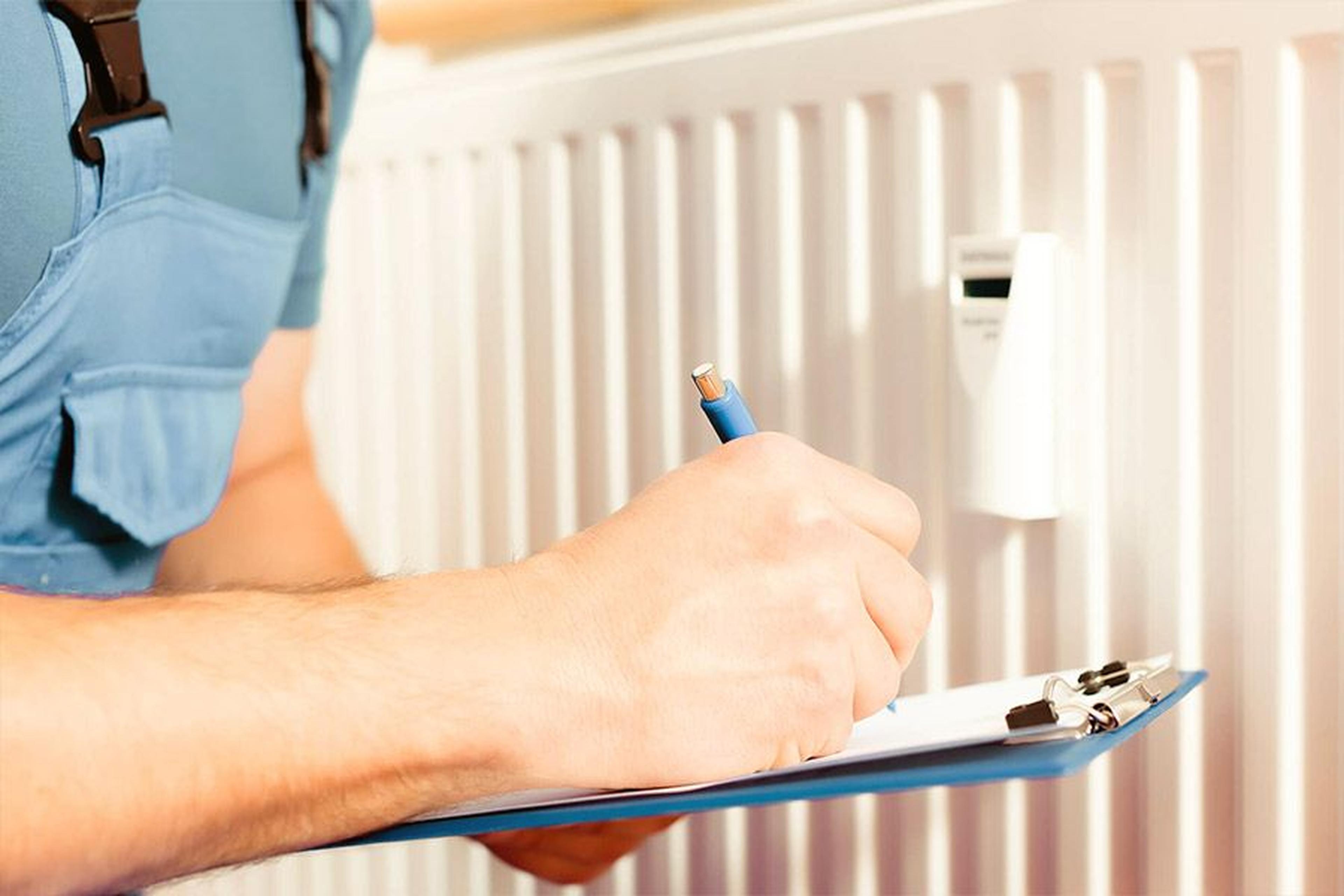 Person in blauer Uniform, die vor einem weißen Heizkörper auf einem Klemmbrett schreibt, möglicherweise führt sie eine Inspektion oder Wartungsprüfung durch.