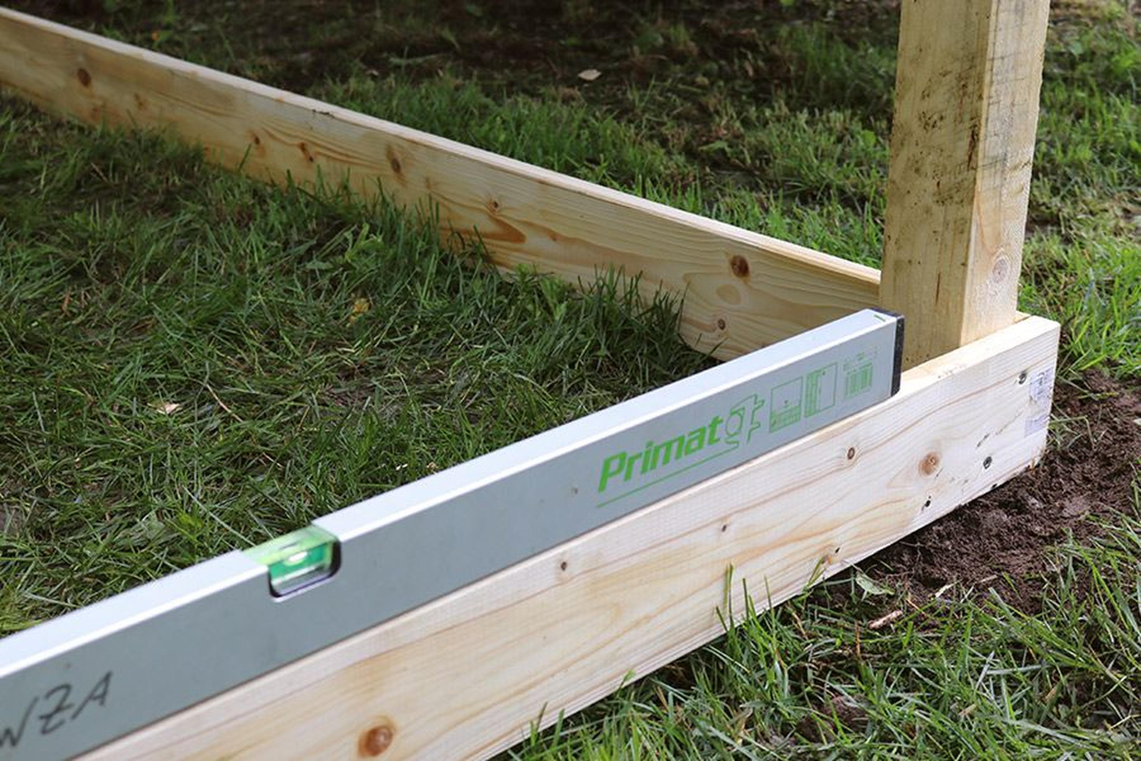 Holzbretter, die das Fundament einer Konstruktion auf Gras bilden, mit einem horizontal platzierten Nivelliergerät zur Ausrichtung.