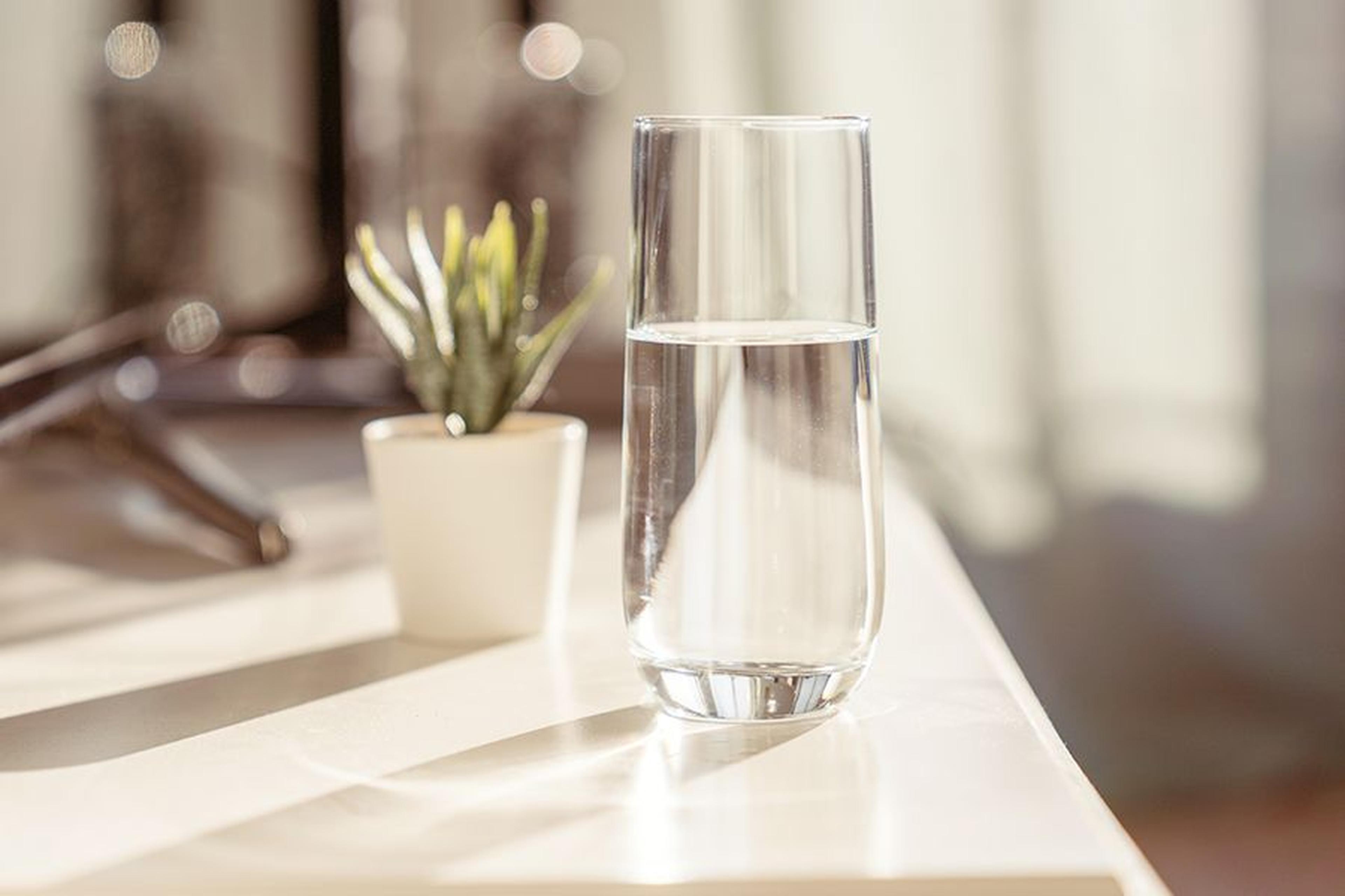 Ein Glas Wasser auf einem sonnenbeschienenen Tisch neben einer kleinen Topfpflanze, mit sanfter Hintergrundbeleuchtung.