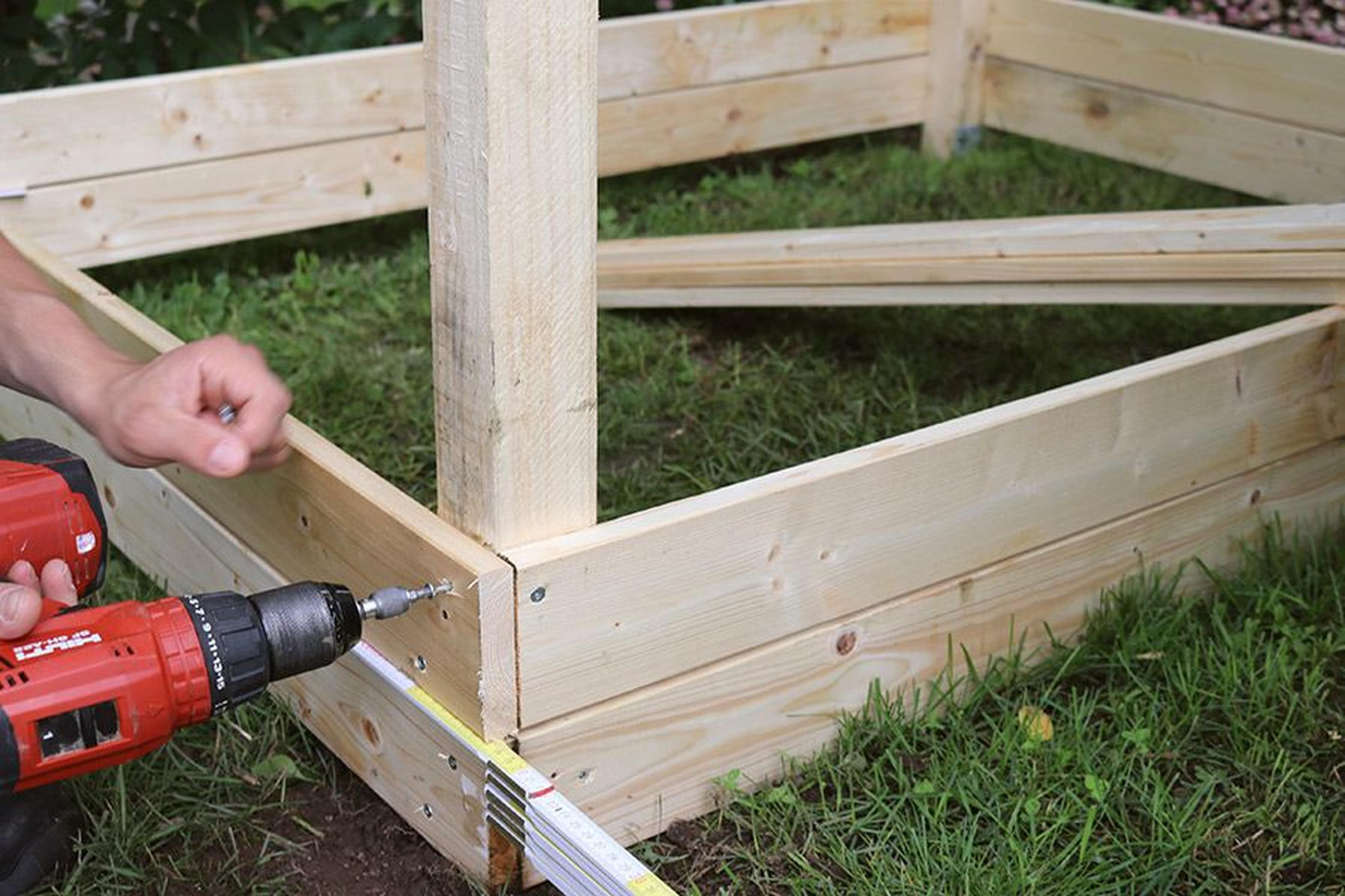 Person, die mit einer roten Bohrmaschine eine Holzkonstruktion auf einer Rasenfläche zusammenbaut, mit einem Maßband in der Nähe.