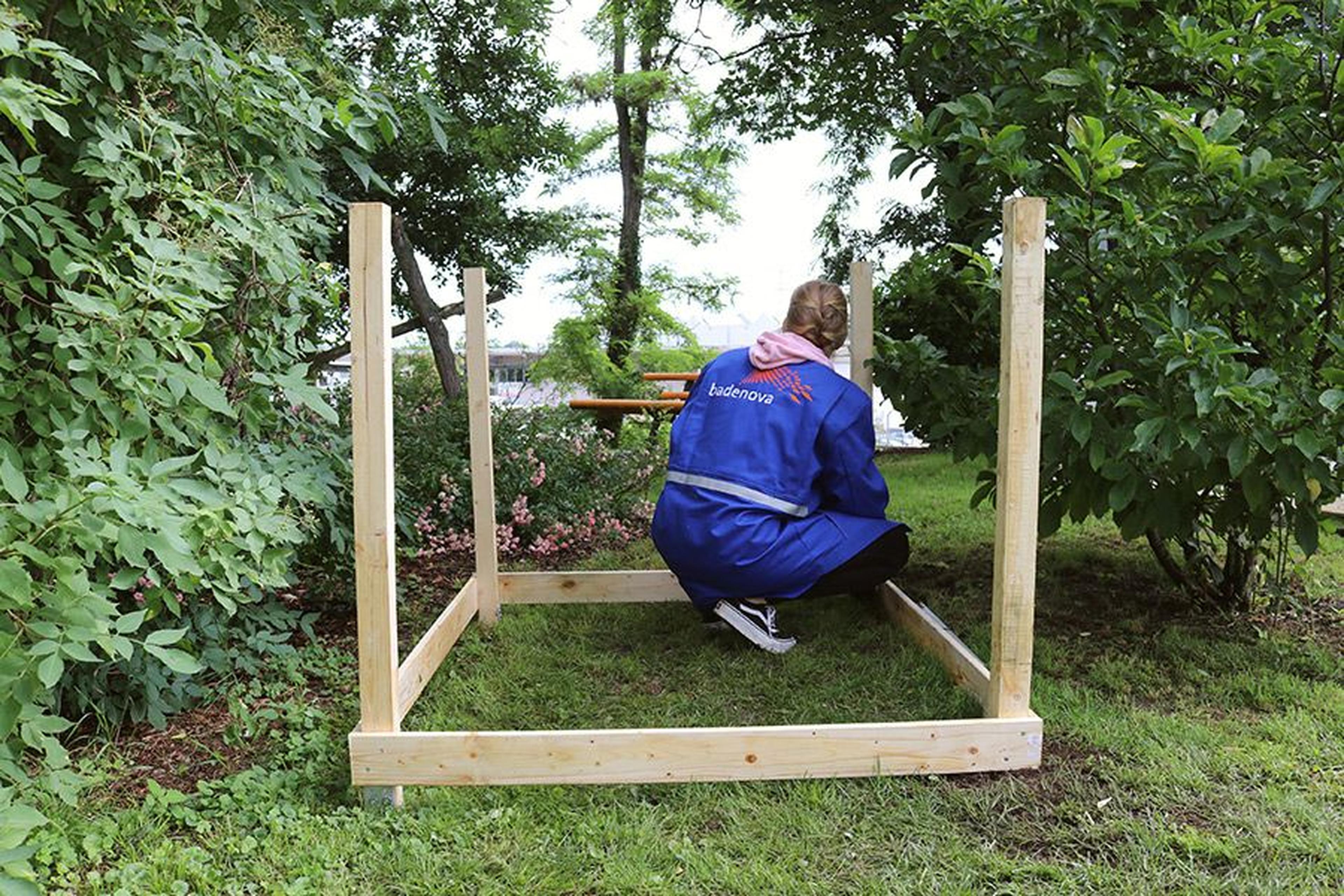 Person in einem blauen Mantel, die draußen einen Holzrahmen zusammenbaut, umgeben von grünen Bäumen und Büschen.