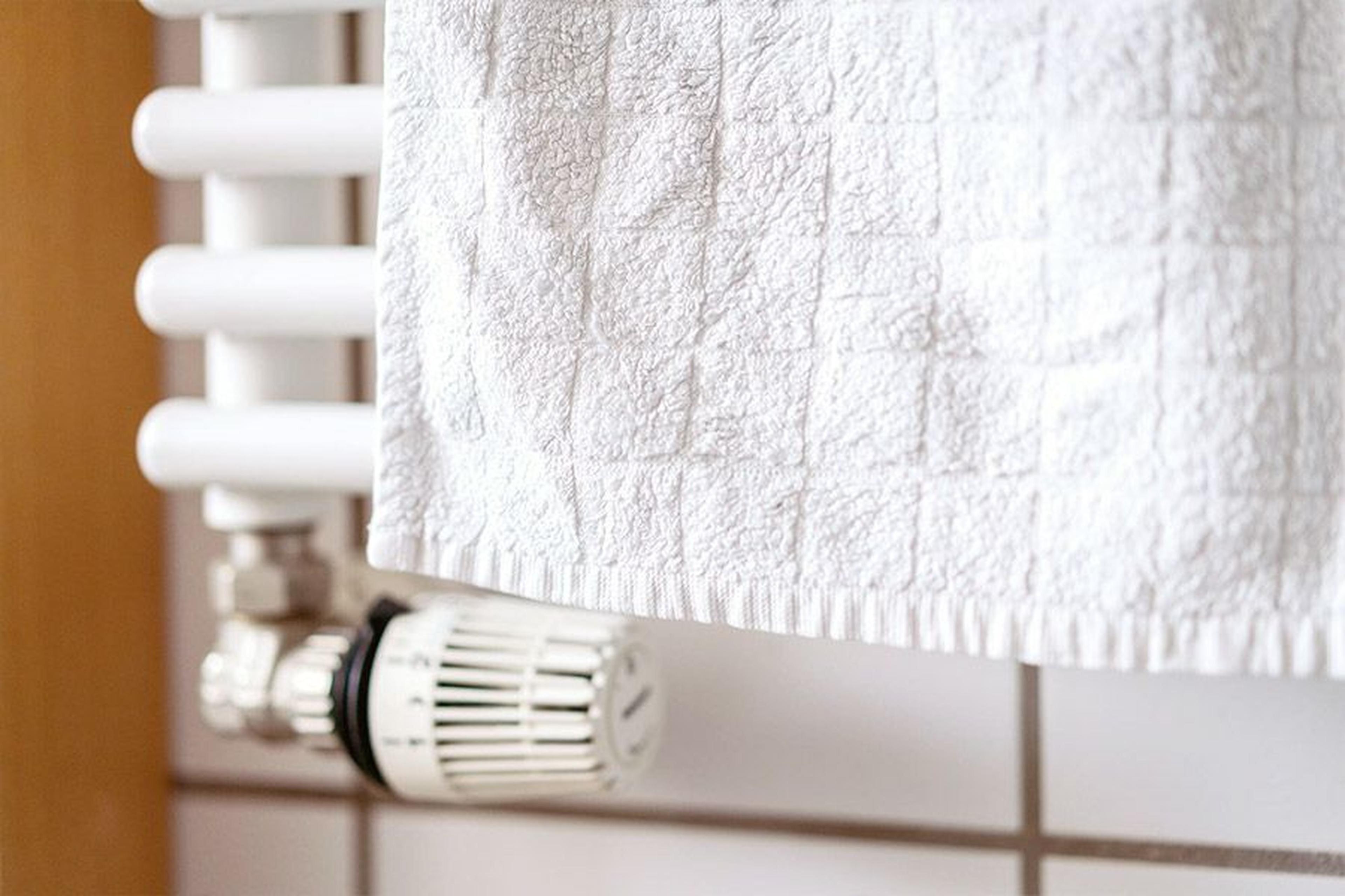 Weißes Handtuch, das an einem beheizten Handtuchhalter mit sichtbarem Thermostatventil hängt, vor einem gefliesten Wandhintergrund.
