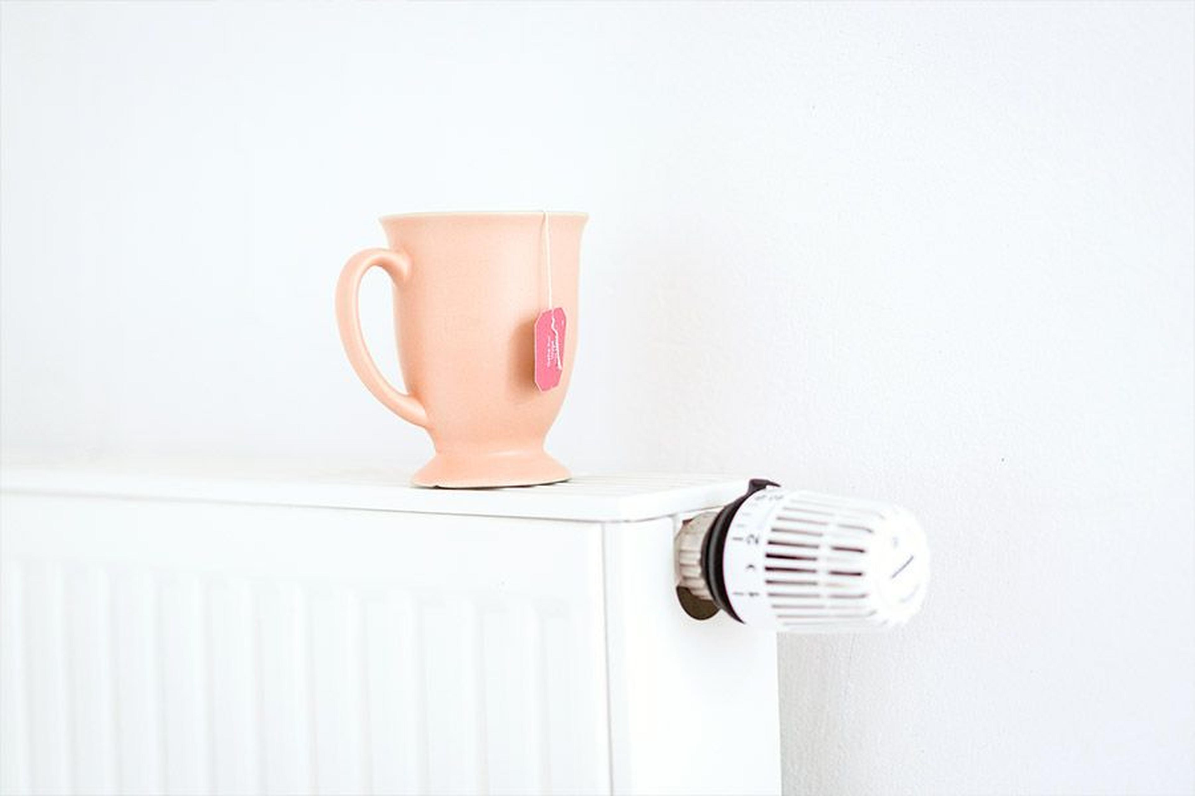 Ein pfirsichfarbener Becher mit einem Teebeutel steht auf einem weißen Heizkörper neben einem Thermostatknopf vor einer schlichten weißen Wand.