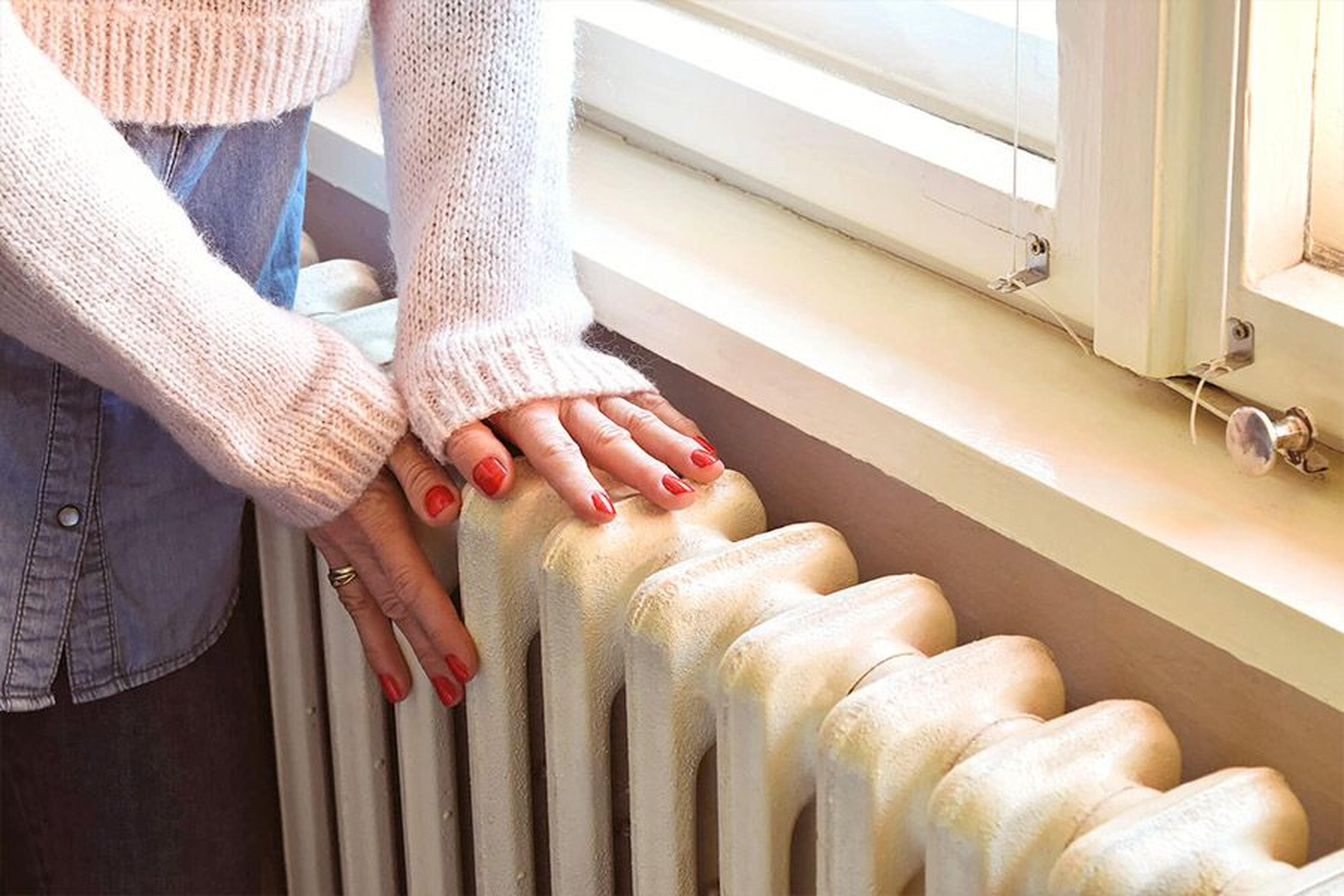Hände mit roten Nägeln ruhen auf einem weißen Heizkörper unter einem Fenster, bekleidet mit einem kuscheligen Pullover.