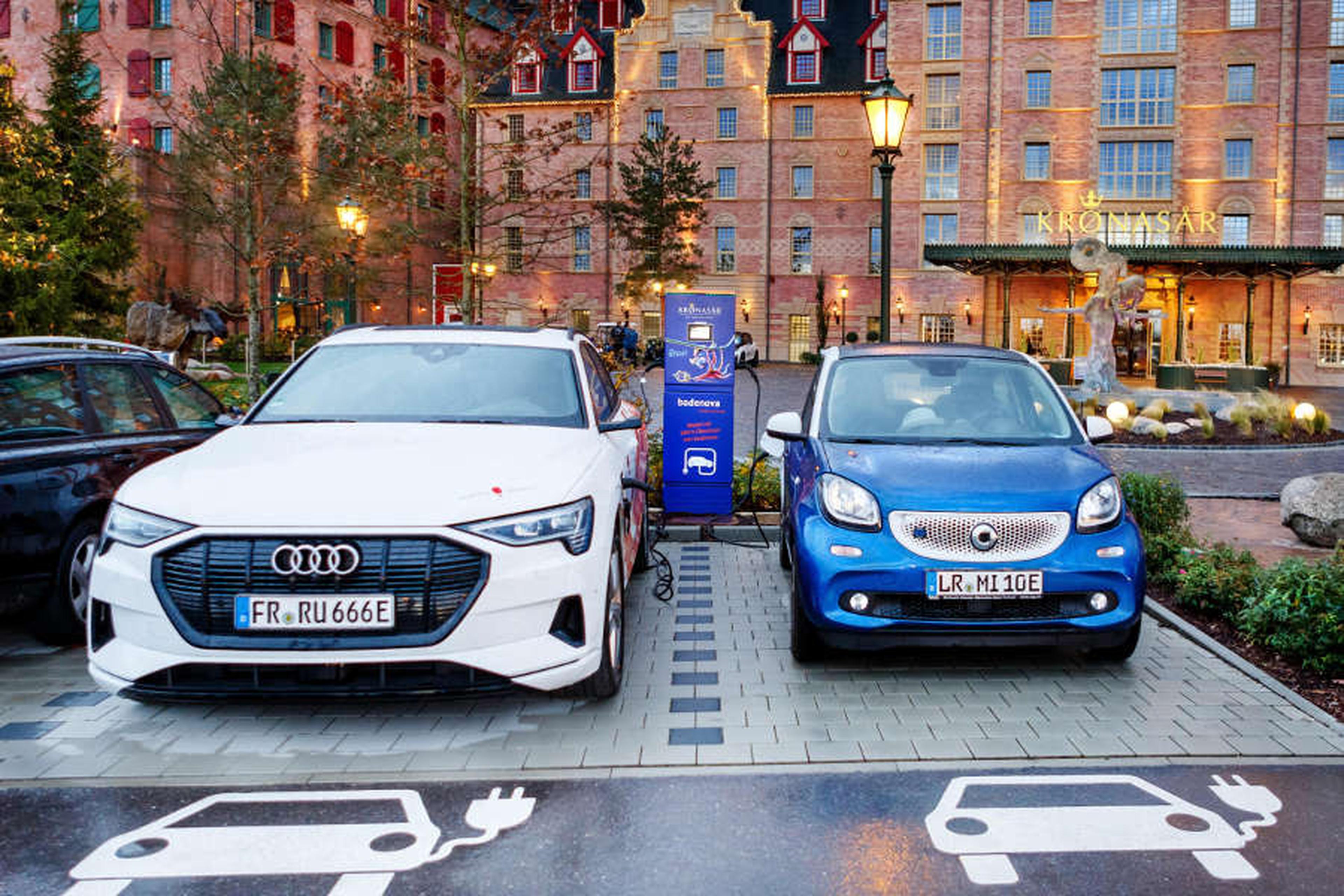 Zwei Elektroautos, ein weißer Audi und ein blauer Smart, laden an einer Station vor einem Backsteingebäude mit dekorativer Beleuchtung.