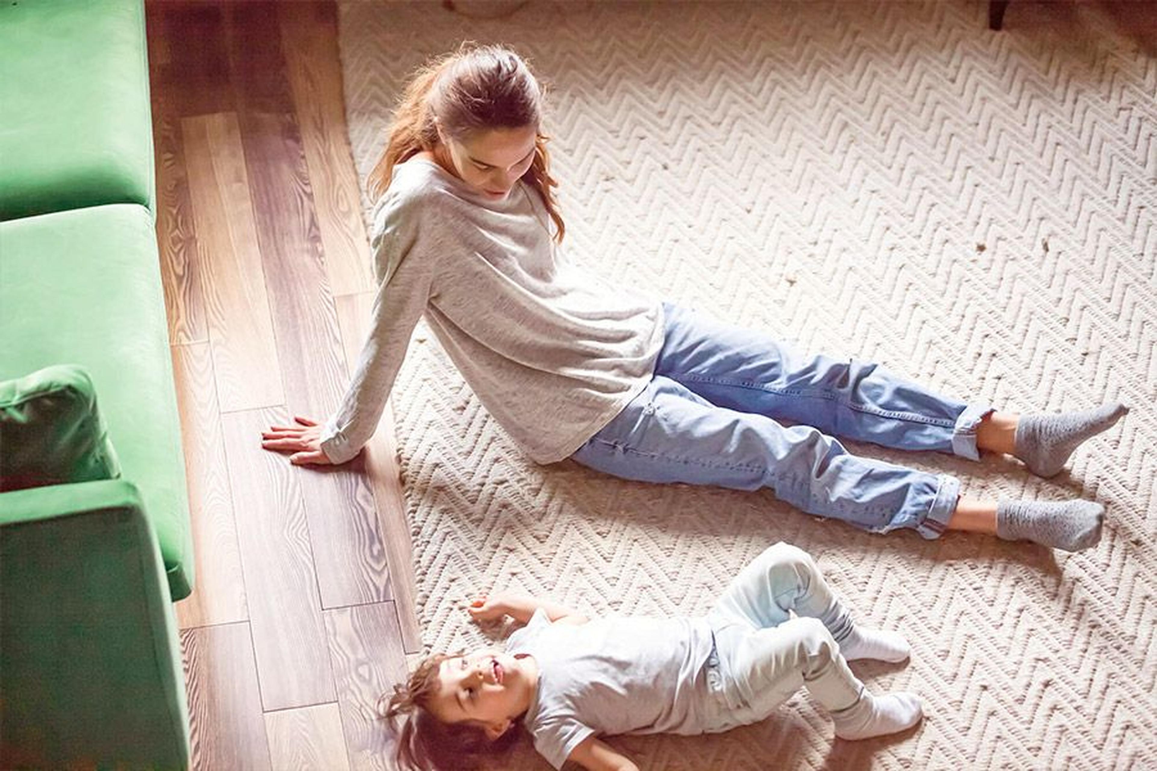 Frau und Kind entspannen sich auf einem gemusterten Teppich, neben ihnen steht ein grünes Sofa. Das Kind liegt auf dem Rücken und schaut zu der sitzenden Frau hinauf.