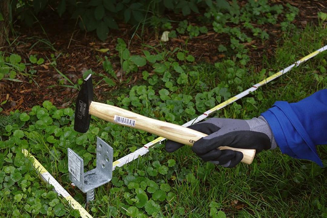 Handschuhhand hält einen Hammer über einem Metallpfahl in grasbewachsenem Boden, umgeben von grünen Blättern und einem gelben Maßband.