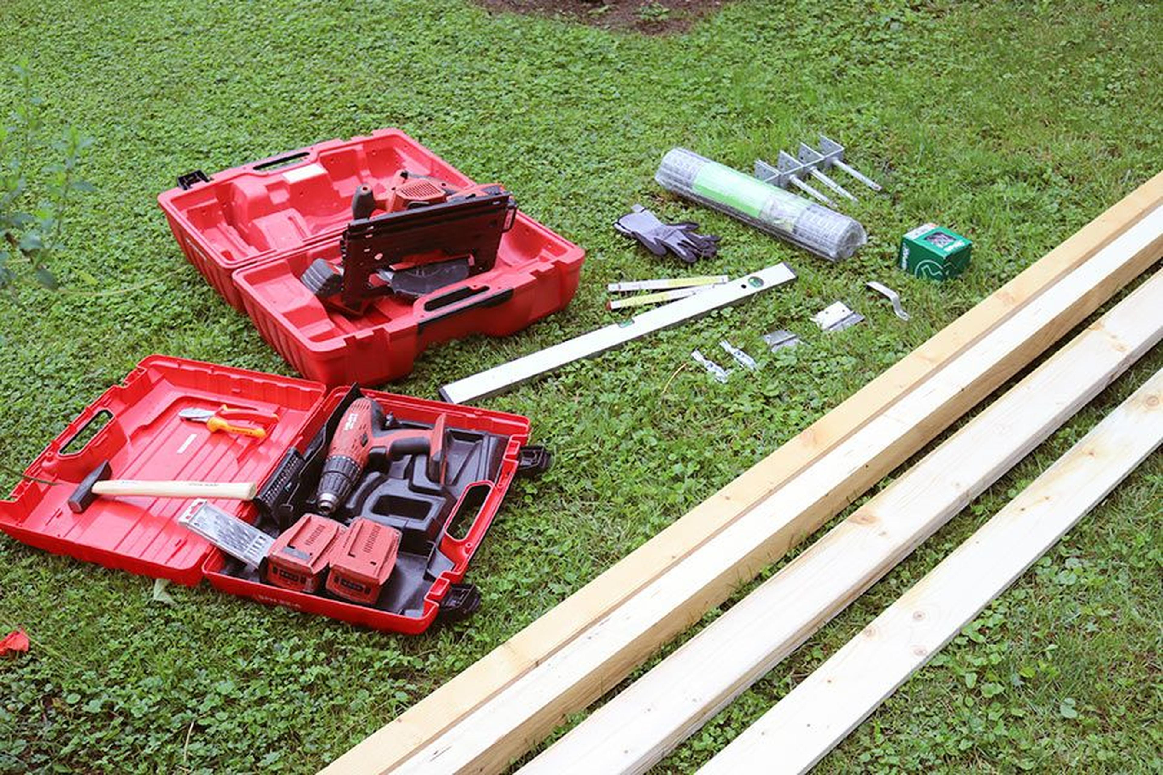 Offene Werkzeugkästen, Holzbretter und Eisenwaren liegen auf dem Rasen verteilt, darunter eine Bohrmaschine, eine Säge, Handschuhe und Nägel.