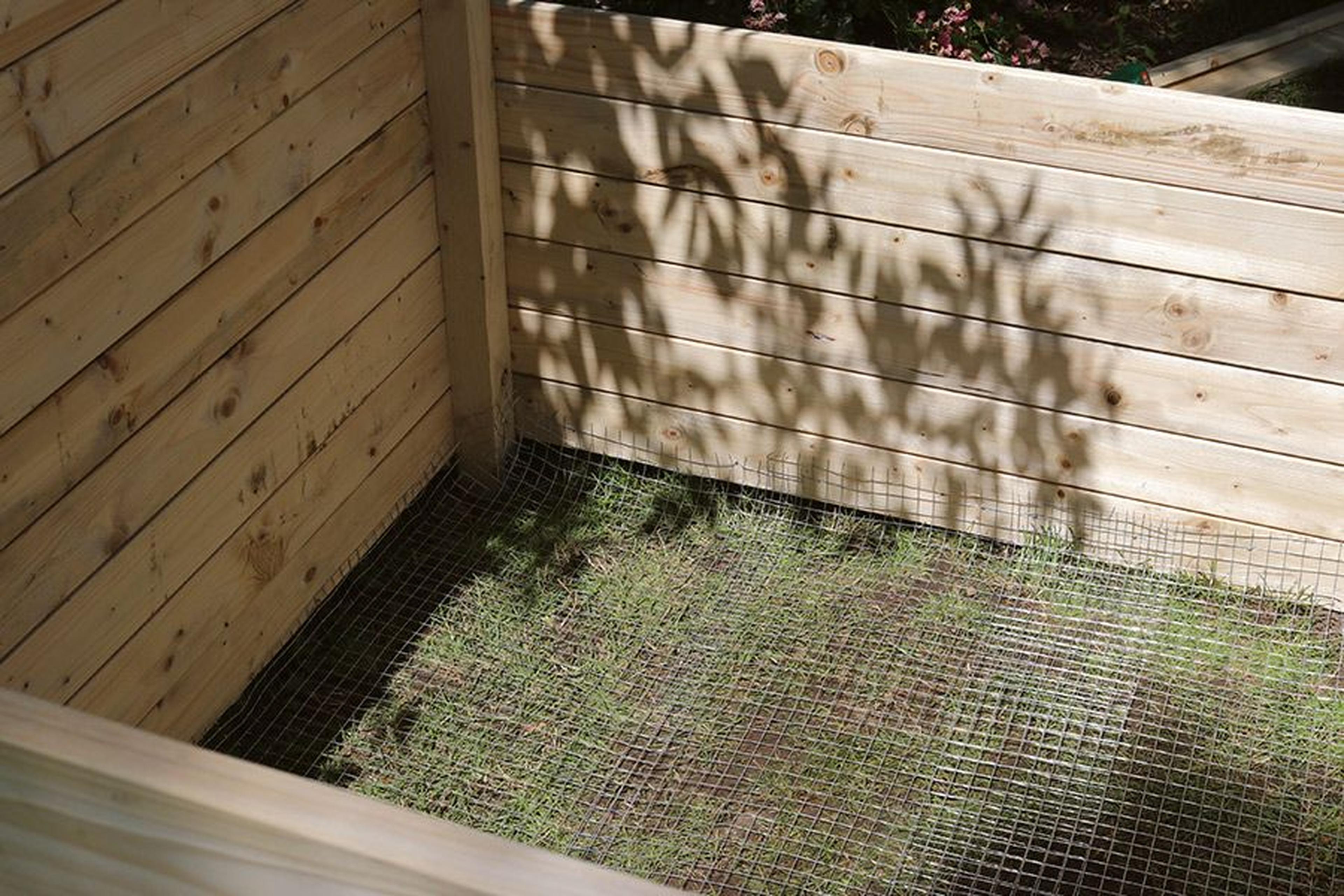 Holzgehege im Garten mit Drahtgitterboden und Gras, teilweise von überhängenden Ästen beschattet.