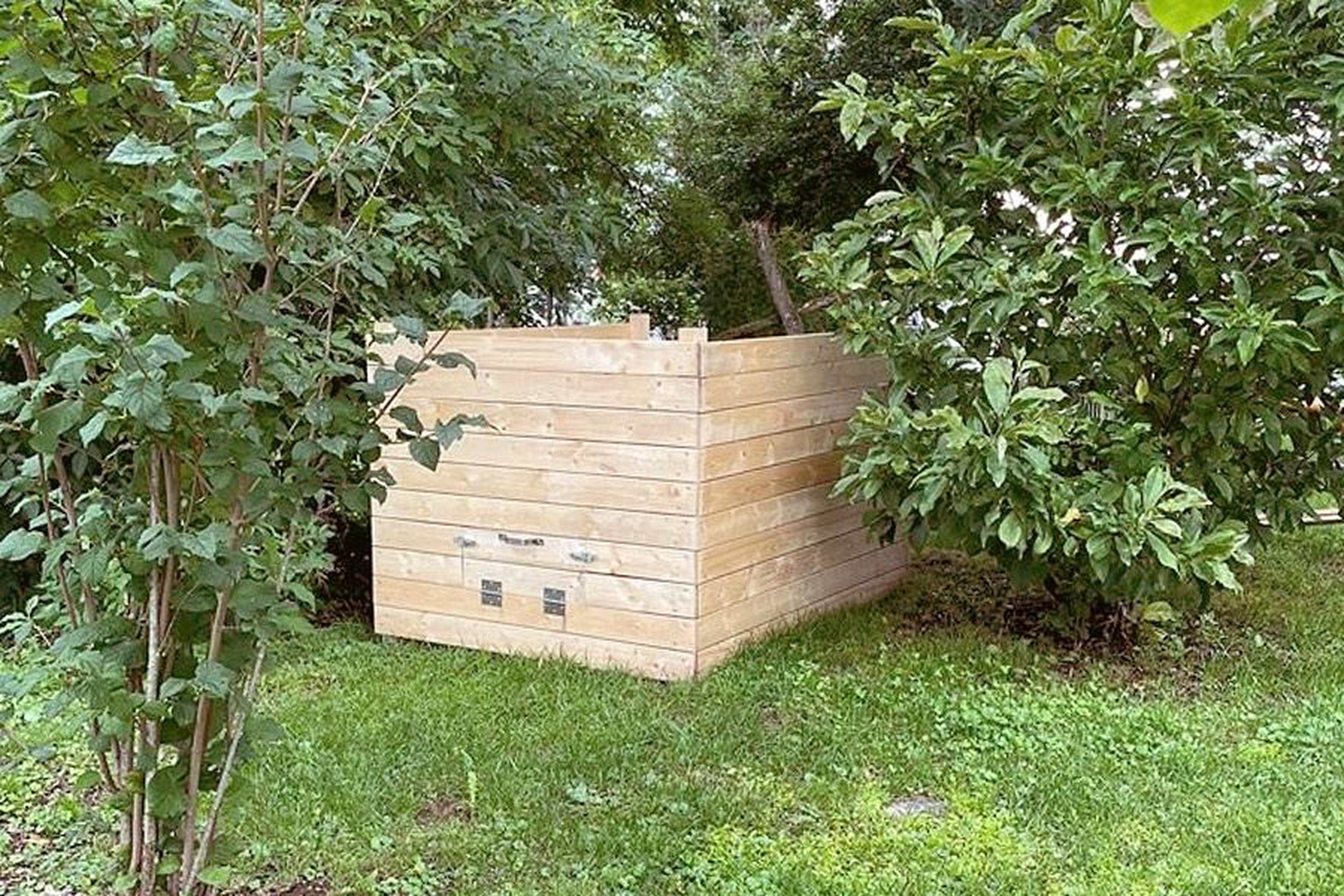 Holzkompostbehälter, umgeben von üppig grünen Bäumen und Gras in einer Gartenumgebung.