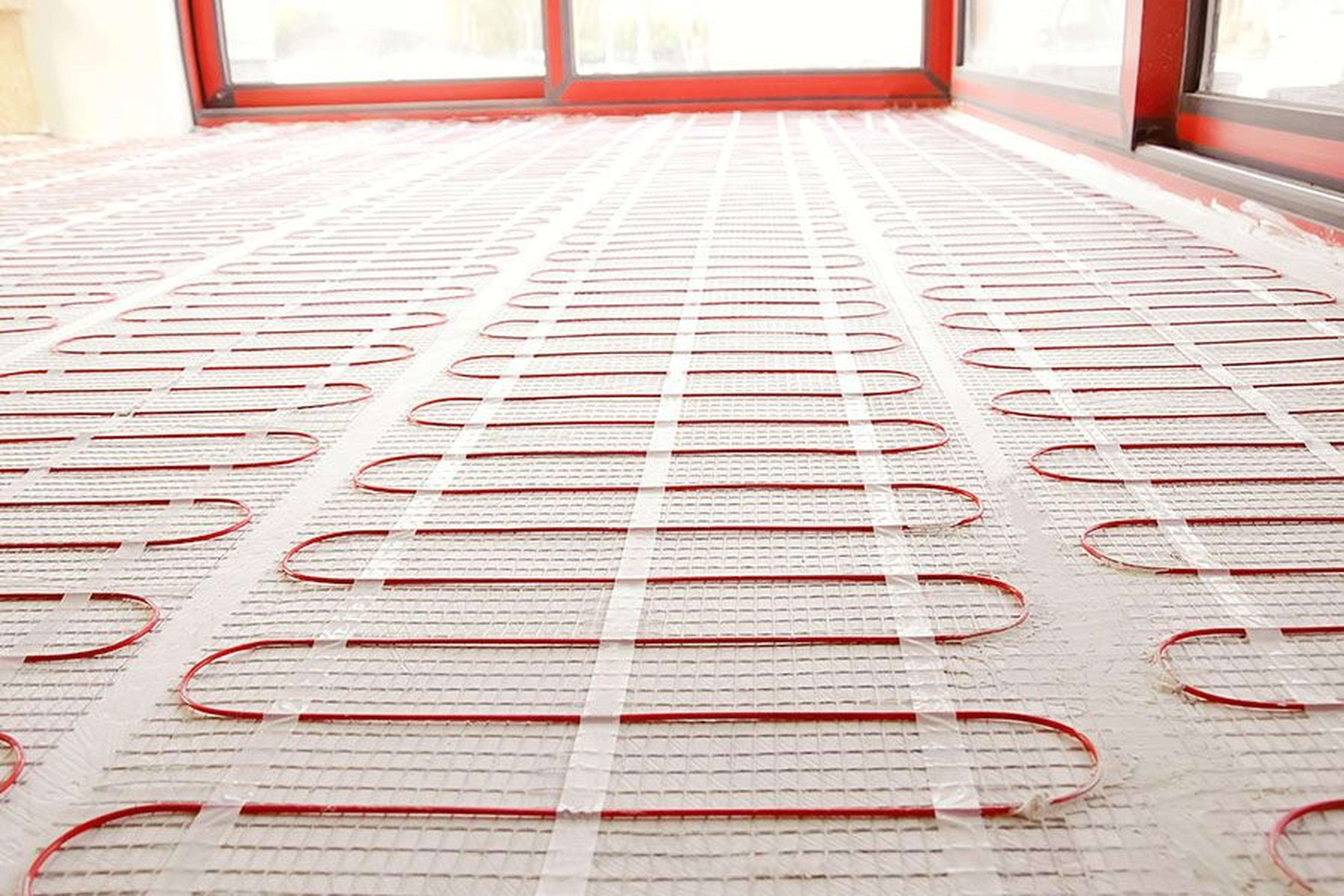 Nahaufnahme einer Fußbodenheizung mit roten Kabeln, die in einem Schleifenmuster auf einem weißen Netz verlegt sind, in der Nähe großer Fenster.
