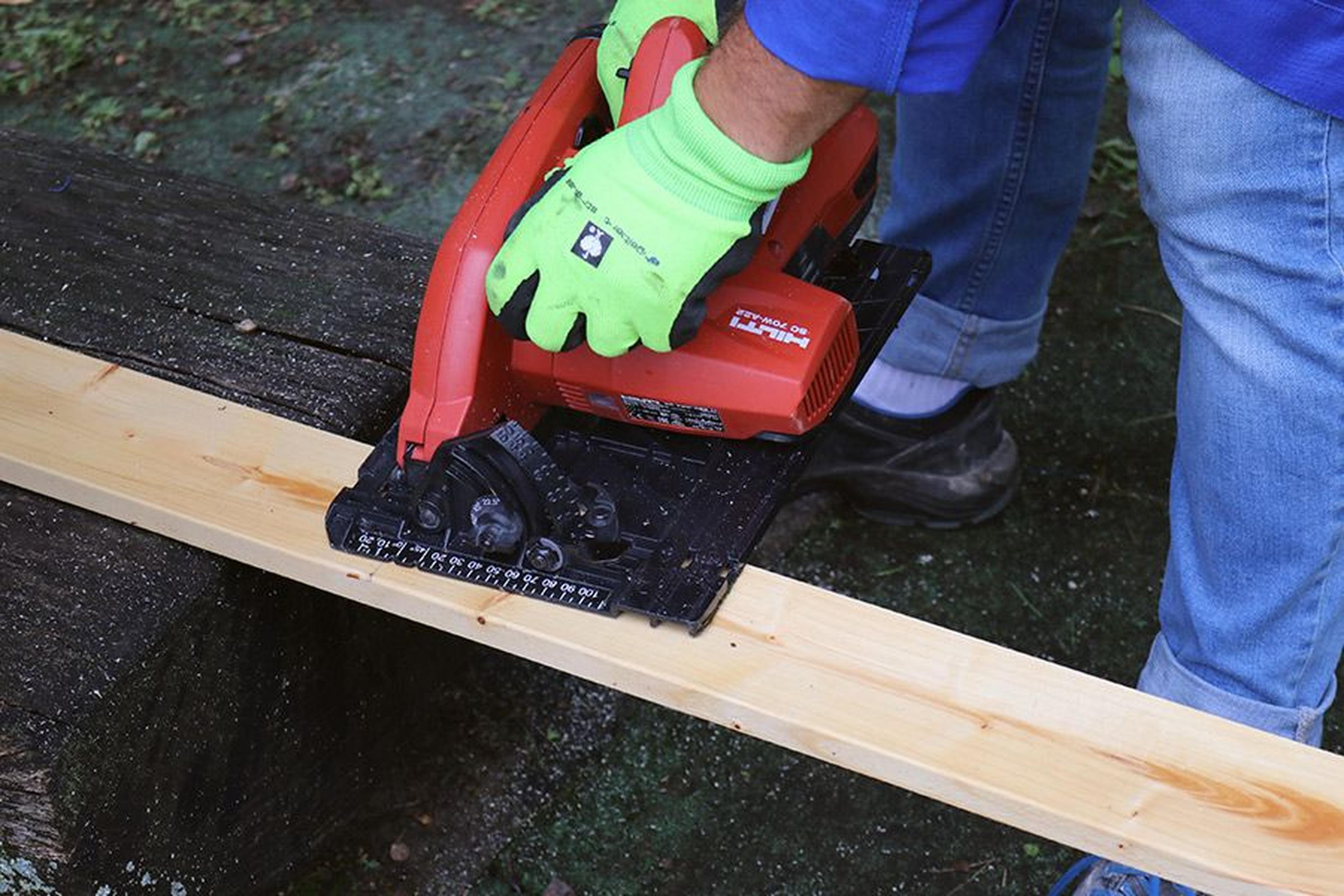 Person mit grünen Handschuhen, die mit einer roten Kreissäge eine Holzplanke auf einer Werkbank im Freien zersägt.