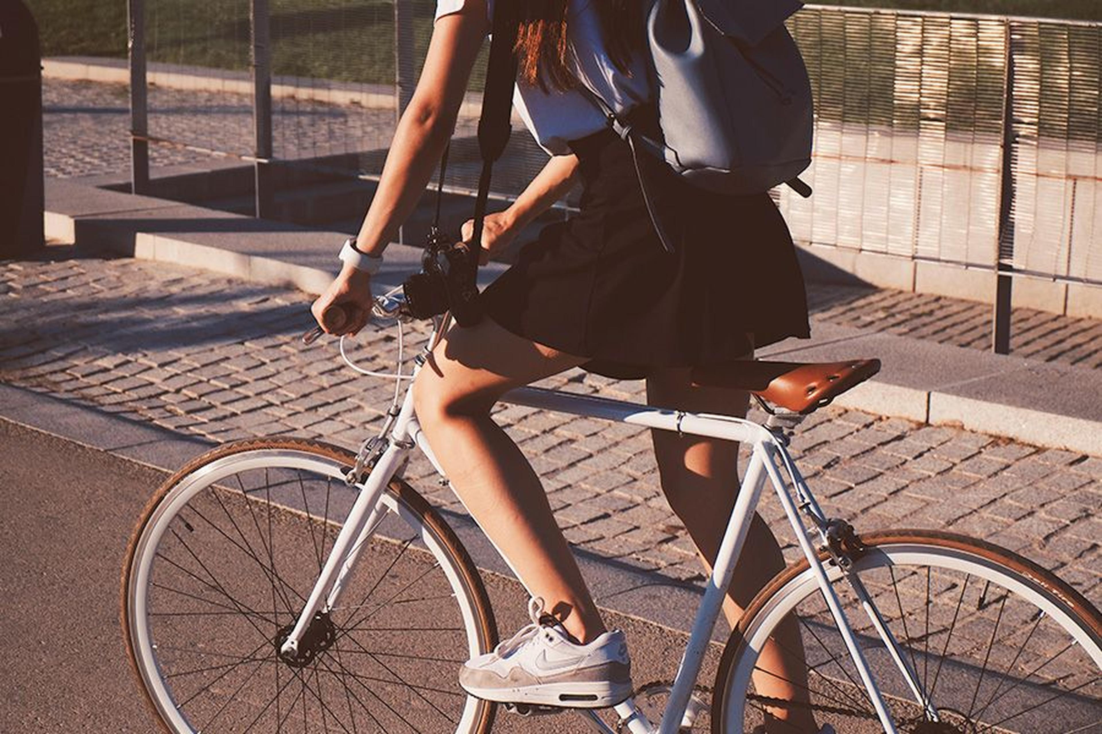Person in einem schwarzen Rock und weißen Turnschuhen, die mit einem blauen Rucksack und einer Kamera auf einem weißen Fahrrad auf einem asphaltierten Weg fährt.