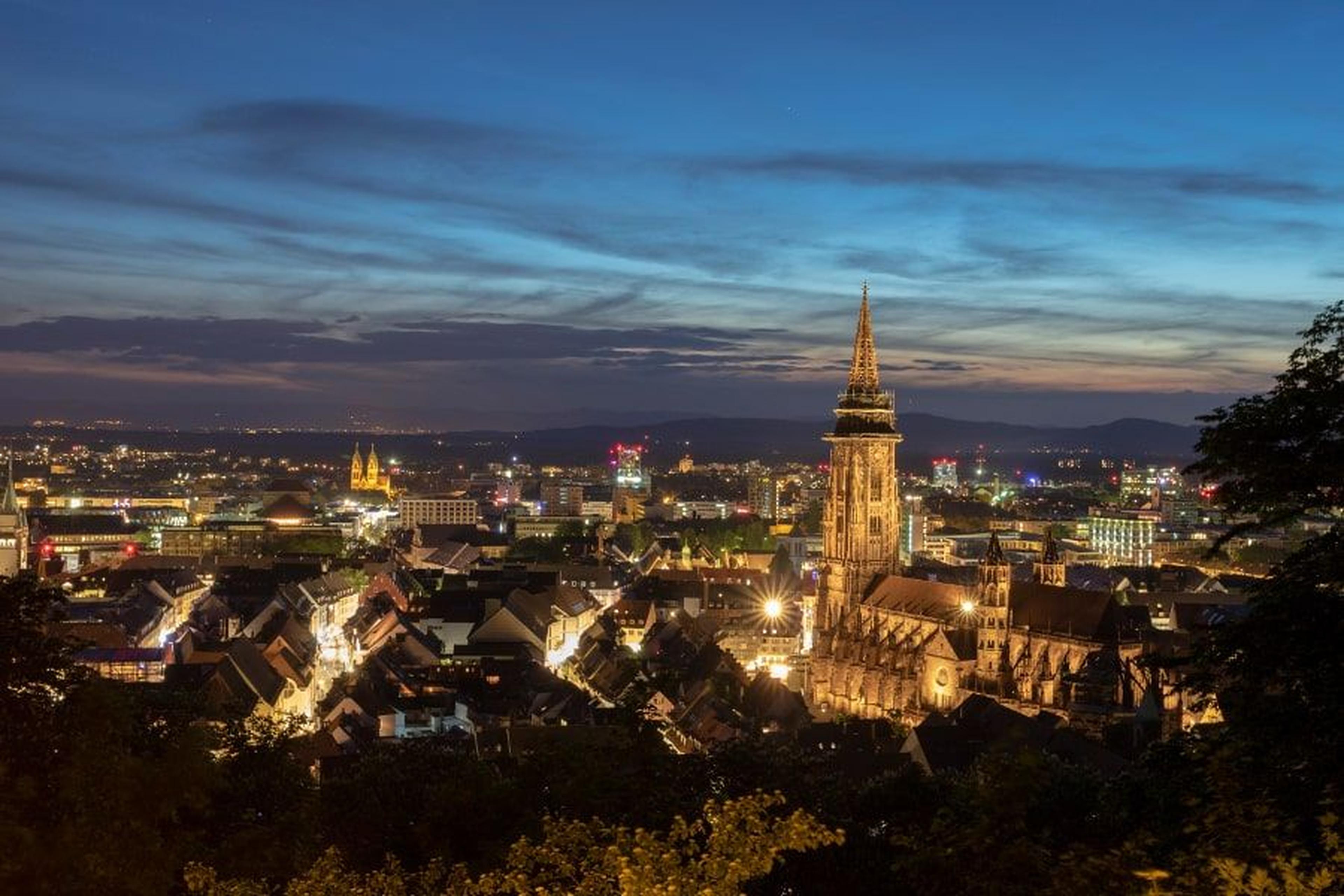 Stadtbild in der Abenddämmerung mit beleuchteten Gebäuden und einem markanten Kirchturm unter einem dämmerigen Himmel mit vereinzelten Wolken.