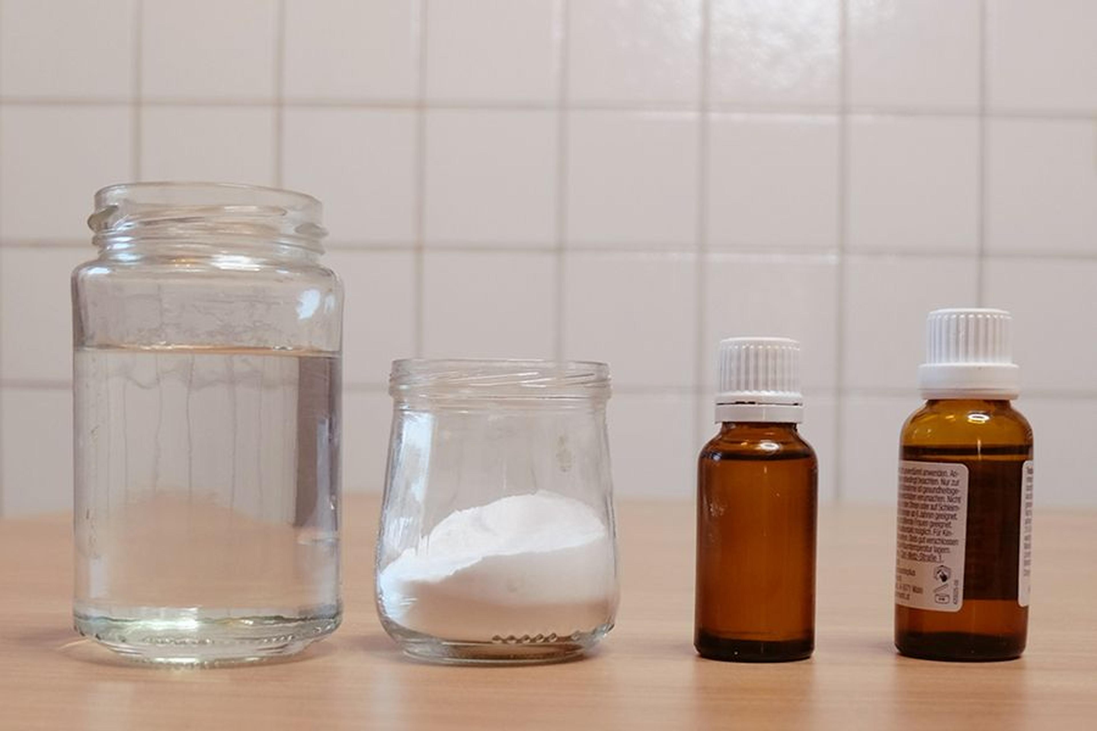 Ein Glas Wasser, ein Glas mit weißem Pulver und zwei bernsteinfarbene Tropfflaschen auf einer Holzoberfläche vor einem gefliesten Hintergrund.