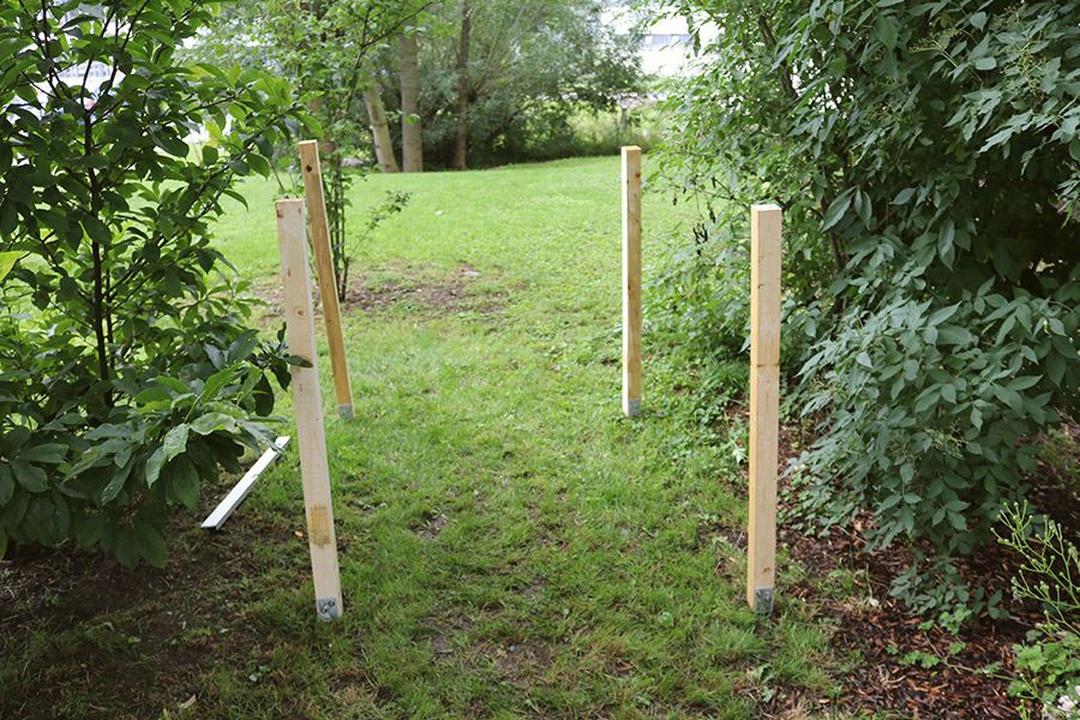 Vier Holzpfosten stehen auf grasbewachsenem Boden und bilden einen rechteckigen Umriss, umgeben von üppigem Grün und Bäumen.