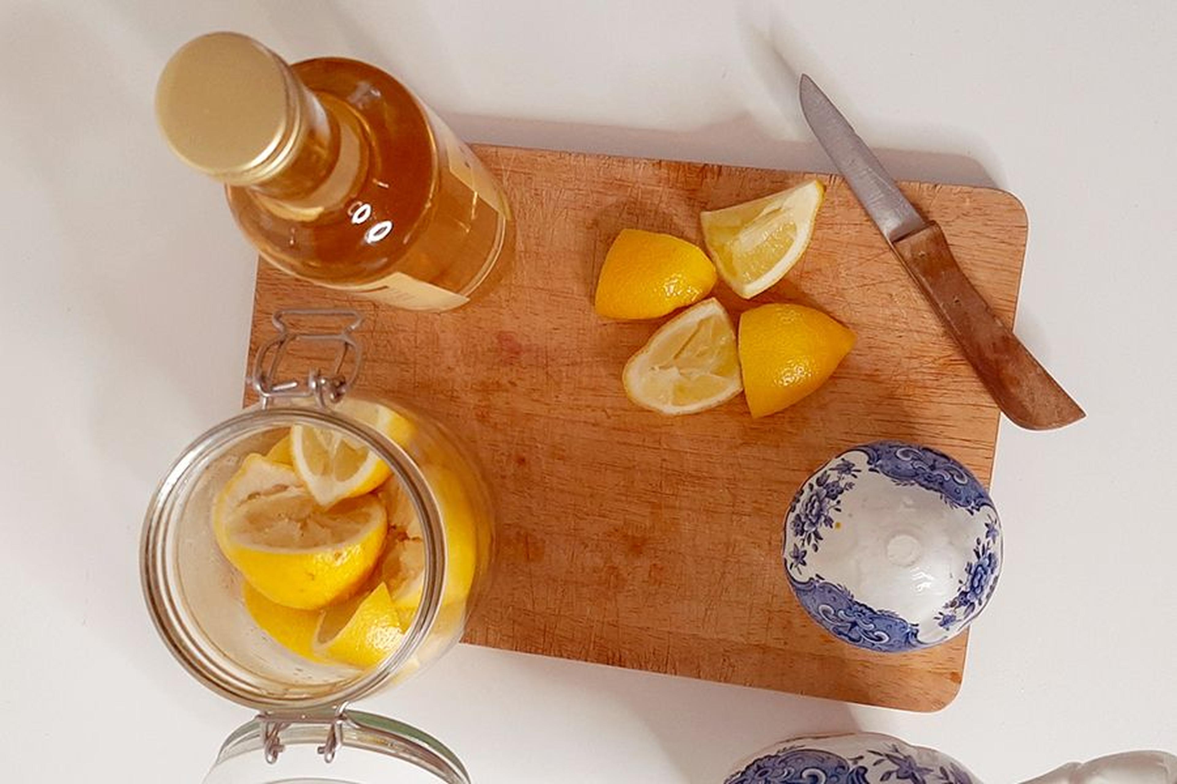 Holzschneidebrett mit geschnittenen Zitronen, einem Messer, einem Glas mit Zitronenscheiben, einer Flasche und einer blau gemusterten Schüssel auf weißem Untergrund.