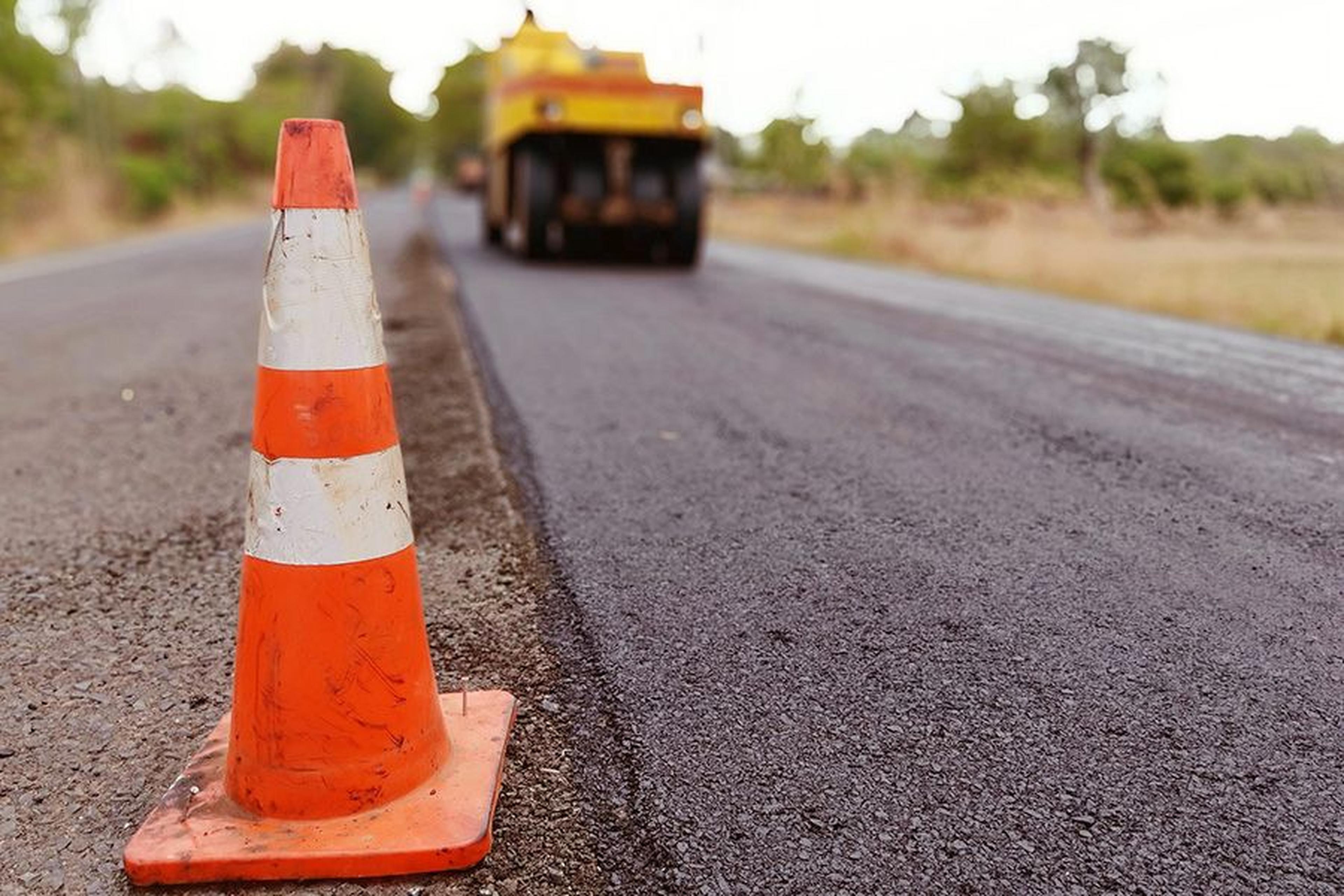 Verkehrsleitkegel steht am Rand einer frischen Asphaltstraße, im Hintergrund ist eine Straßenbaumaschine unscharf zu sehen.