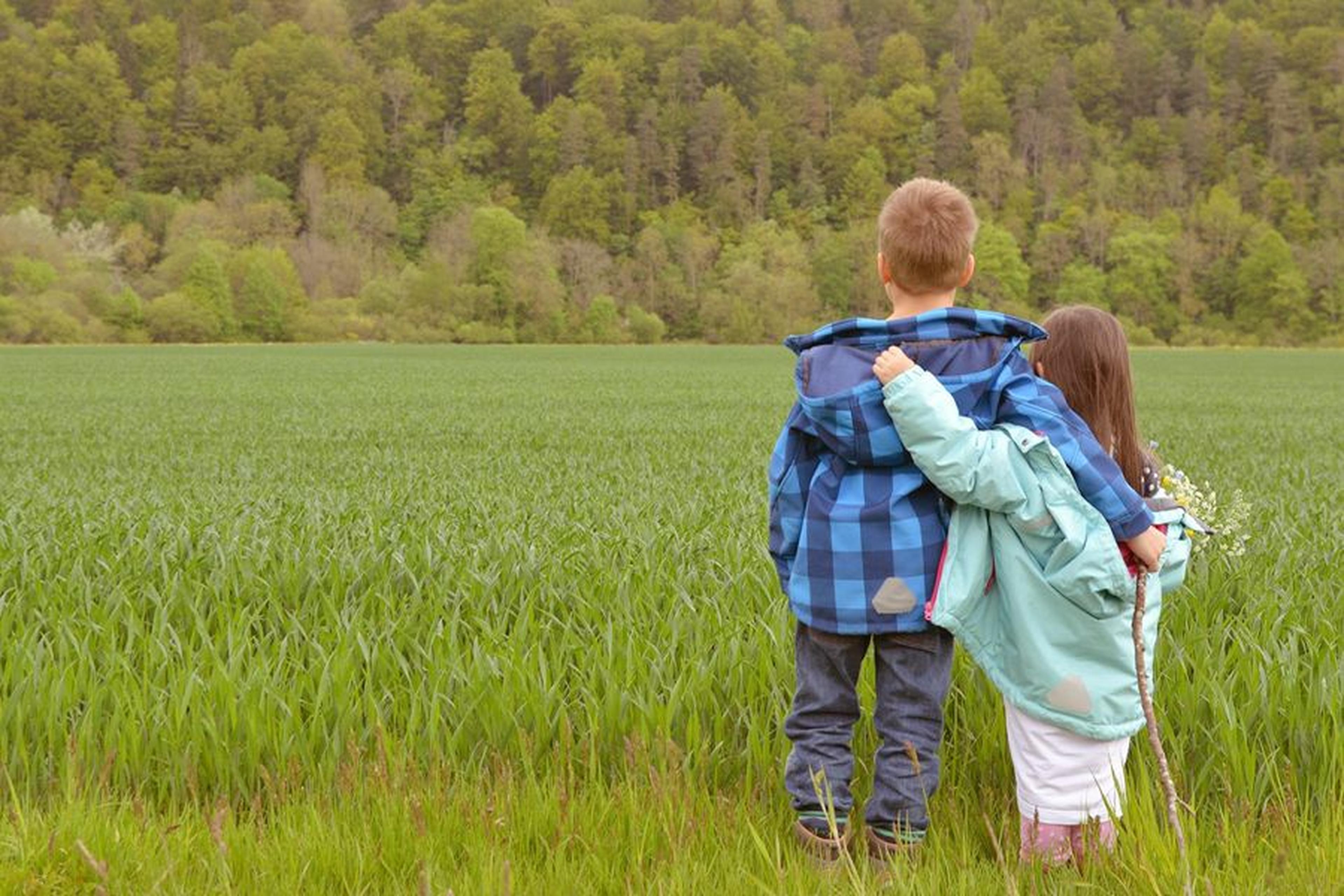 Zwei Kinder in Jacken stehen sich auf einer grünen Wiese umarmt gegenüber, im Hintergrund ist ein Wald zu sehen. Eines der Kinder hält einen kleinen Blumenstrauß in der Hand.
