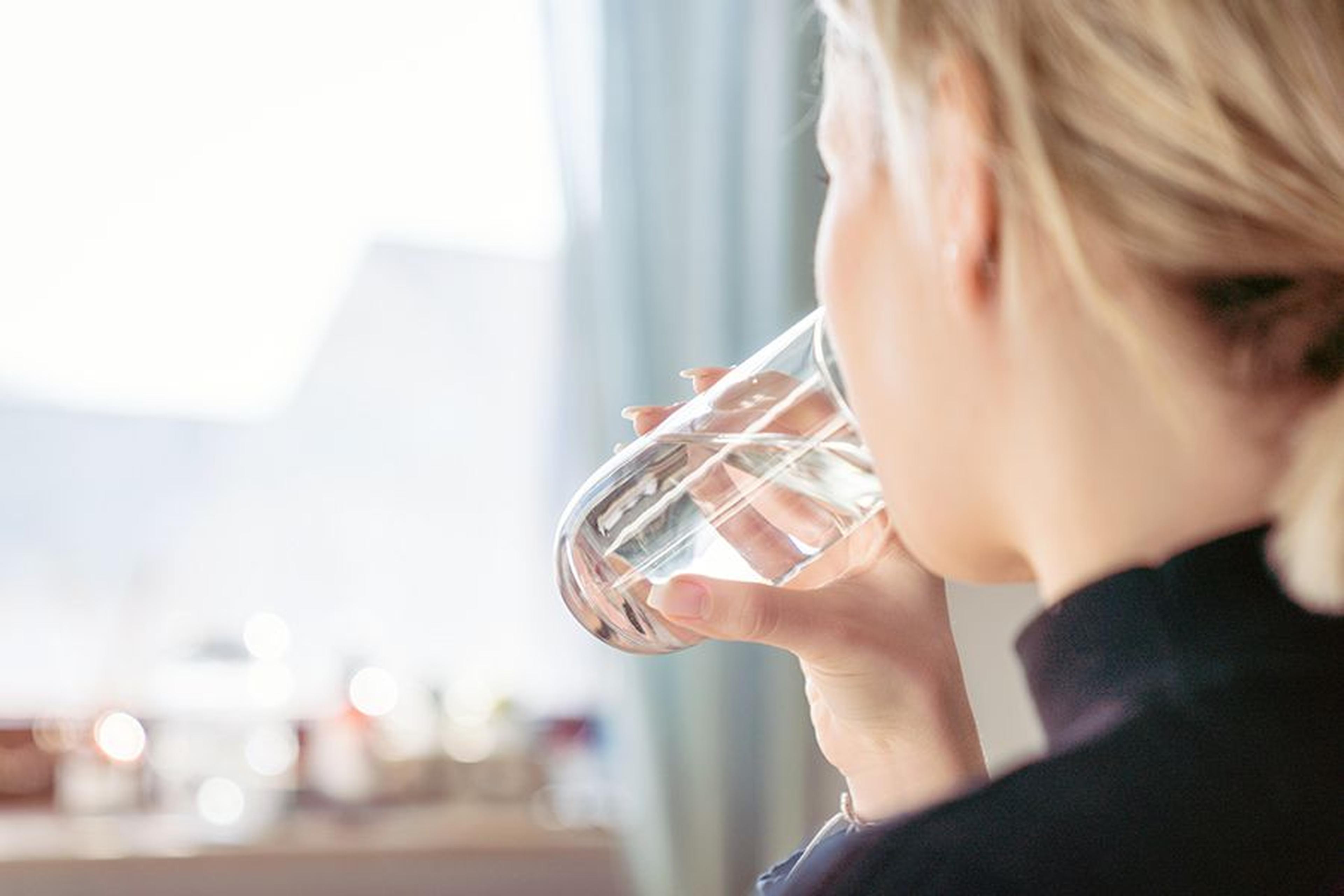 Eine Person mit blonden Haaren trinkt ein Glas Wasser und sitzt dabei in der Nähe eines sonnendurchfluteten Fensters.