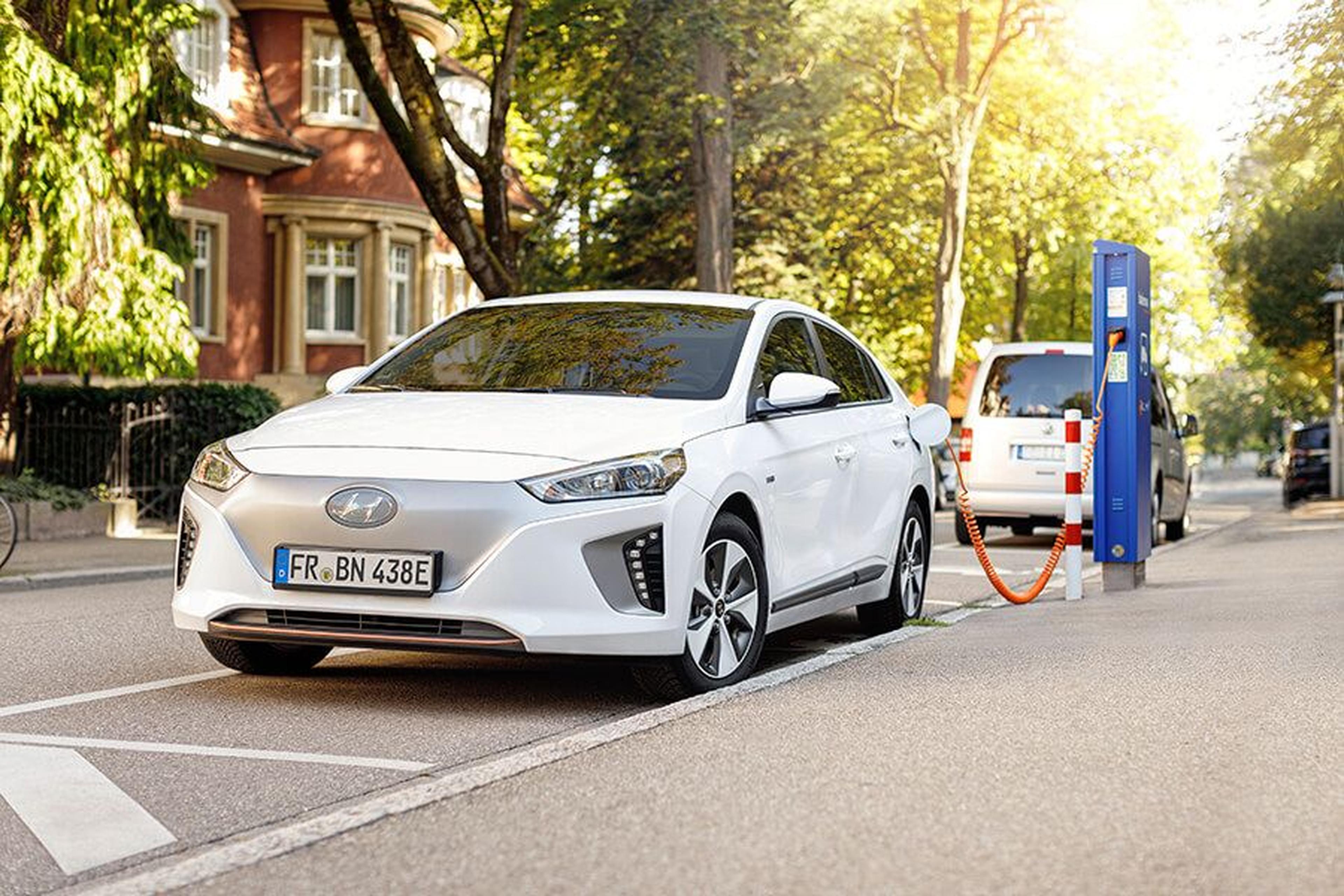 Weißes Elektroauto, das an einer Ladestation am Straßenrand in einem grünen Stadtgebiet aufgeladen wird, während Sonnenlicht durch die Bäume scheint.