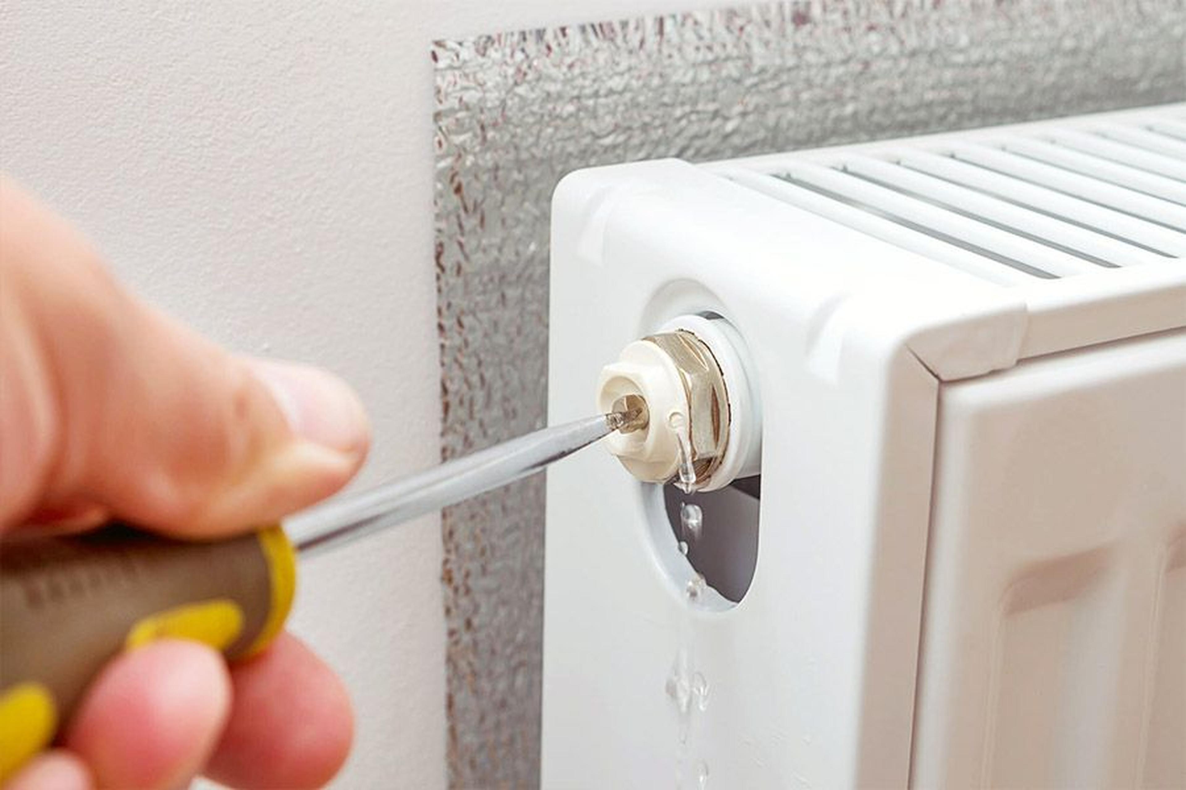 Nahaufnahme einer Hand, die mit einem Schraubendreher einen Heizkörper entlüftet, wobei Wasser aus dem Ventil tropft.