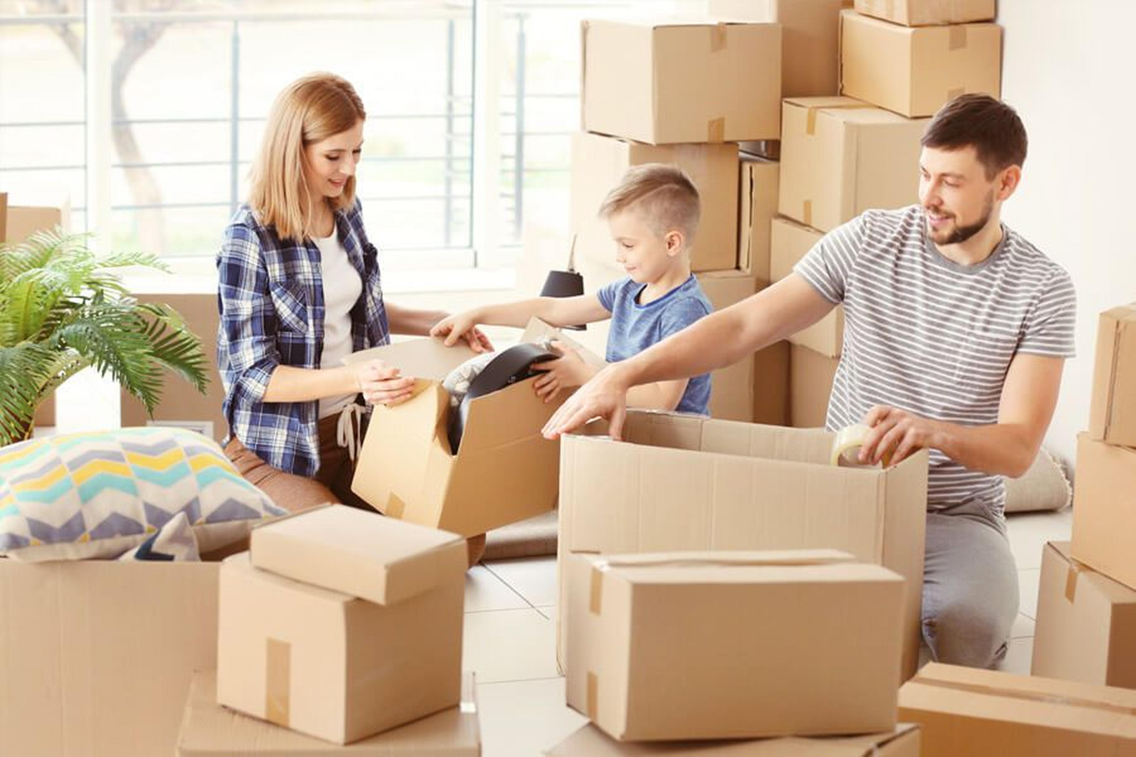 Eine Familie packt in einem hellen Raum Kisten und bereitet sich auf den Umzug vor. Eine Frau, ein Kind und ein Mann sortieren Gegenstände zwischen gestapelten Kartons.