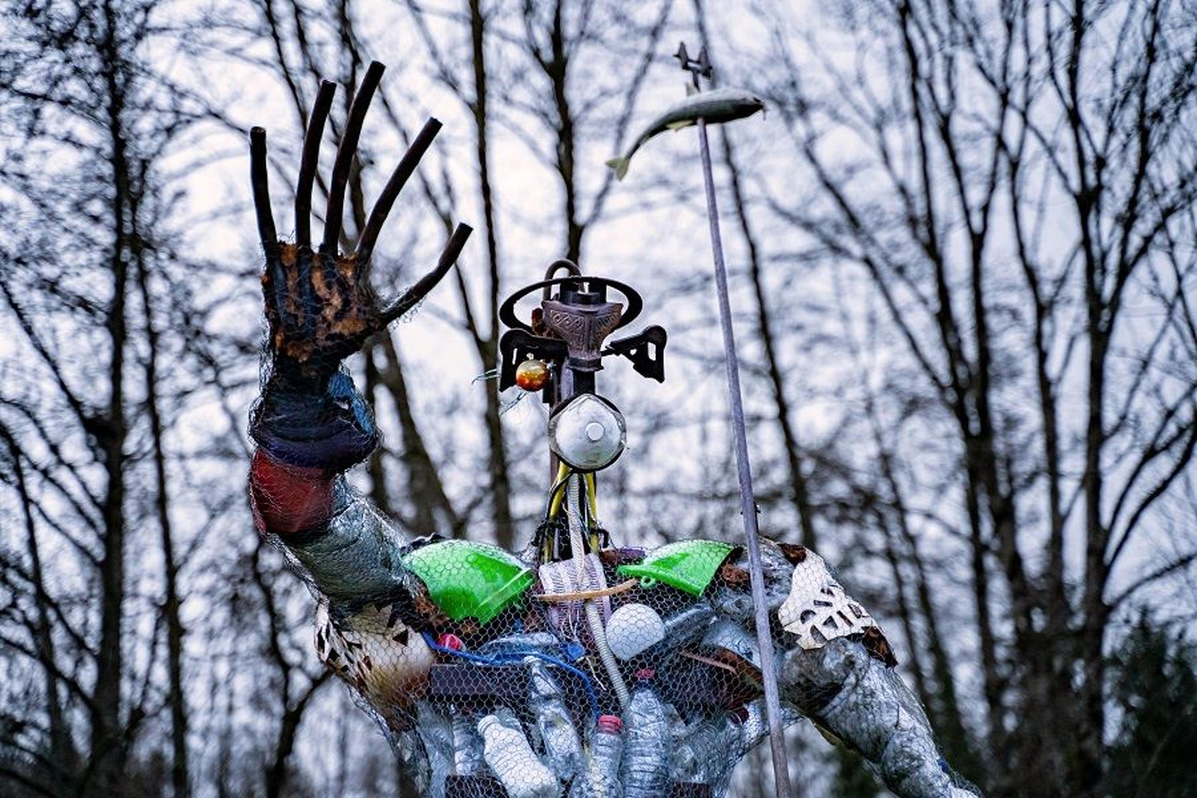 Skulptur in Menschengestalt, zusammengesetzt aus Plastikflaschen und Abfällen, steht im Freien zwischen Bäumen.