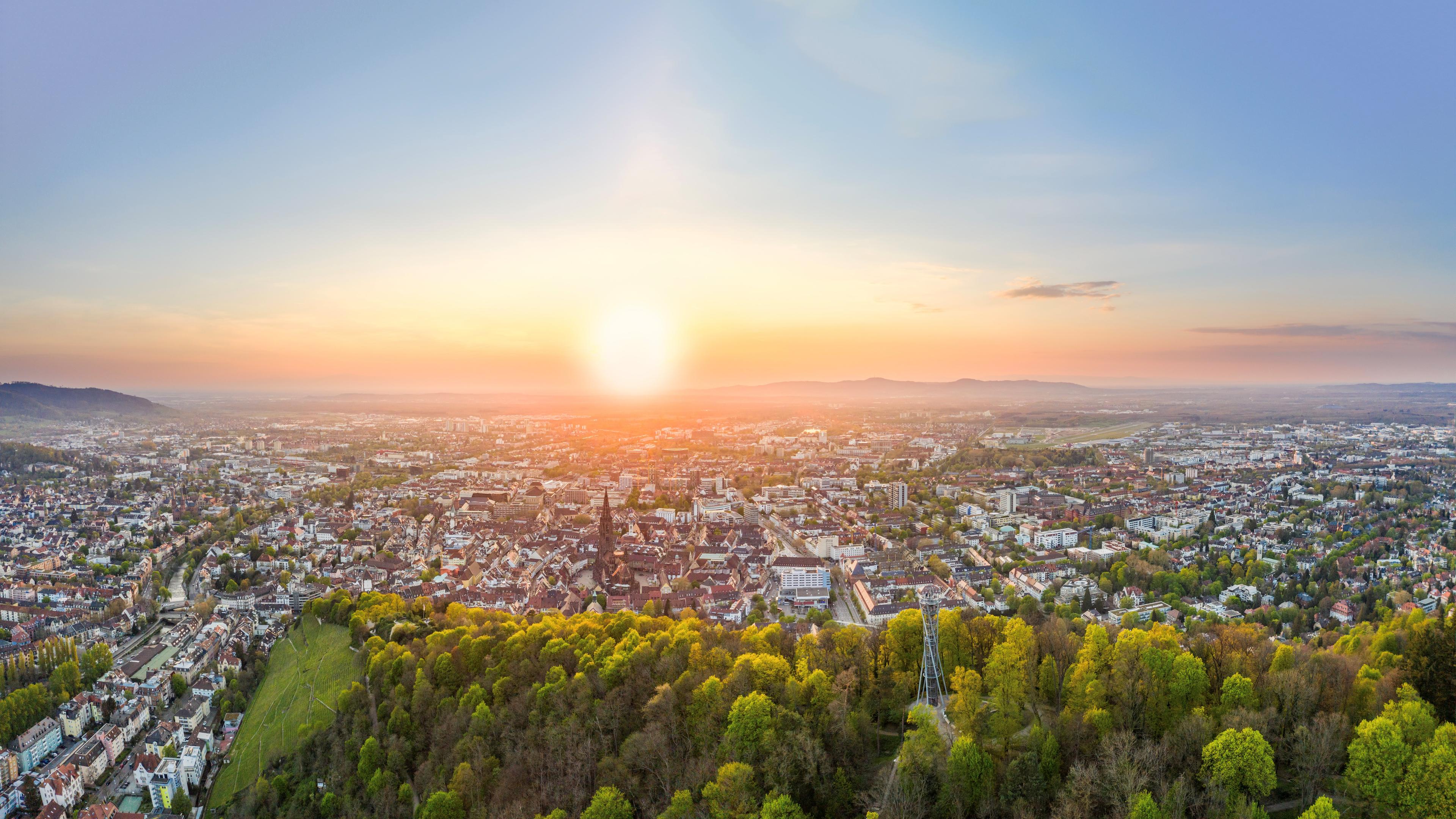 anoramaaufnahme einer Stadt bei Sonnenuntergang, mit dicht bebauten Stadtbereichen im Vordergrund und grünen Baumflächen am unteren Bildrand. Die Sonne steht tief am Horizont und taucht die Szene in warmes Licht, während im Hintergrund Hügel und Berge sichtbar sind
