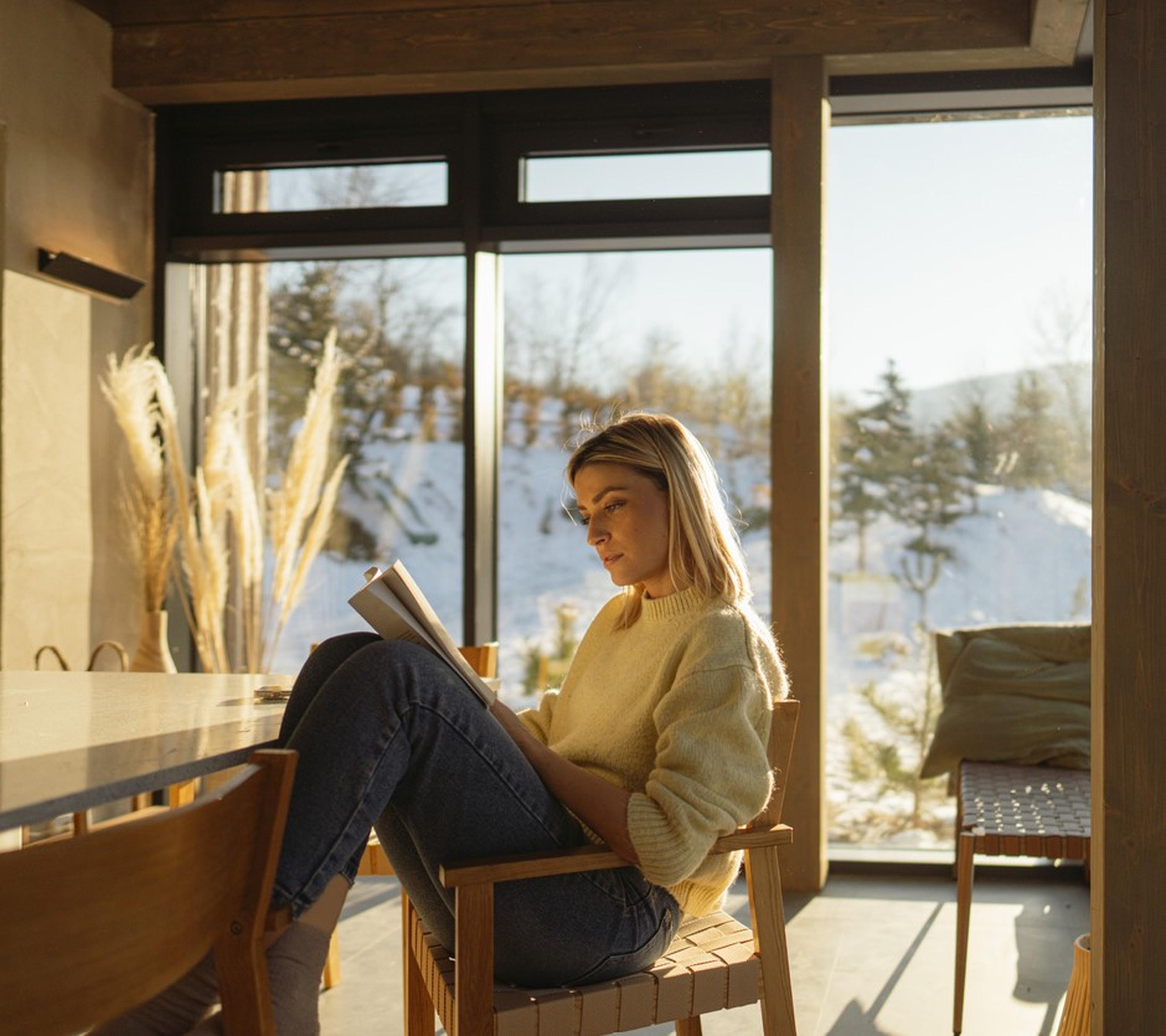 Frau sitzt lesend am Tisch, während draußen Schnee liegt und die Sonne durchs Fenster scheint