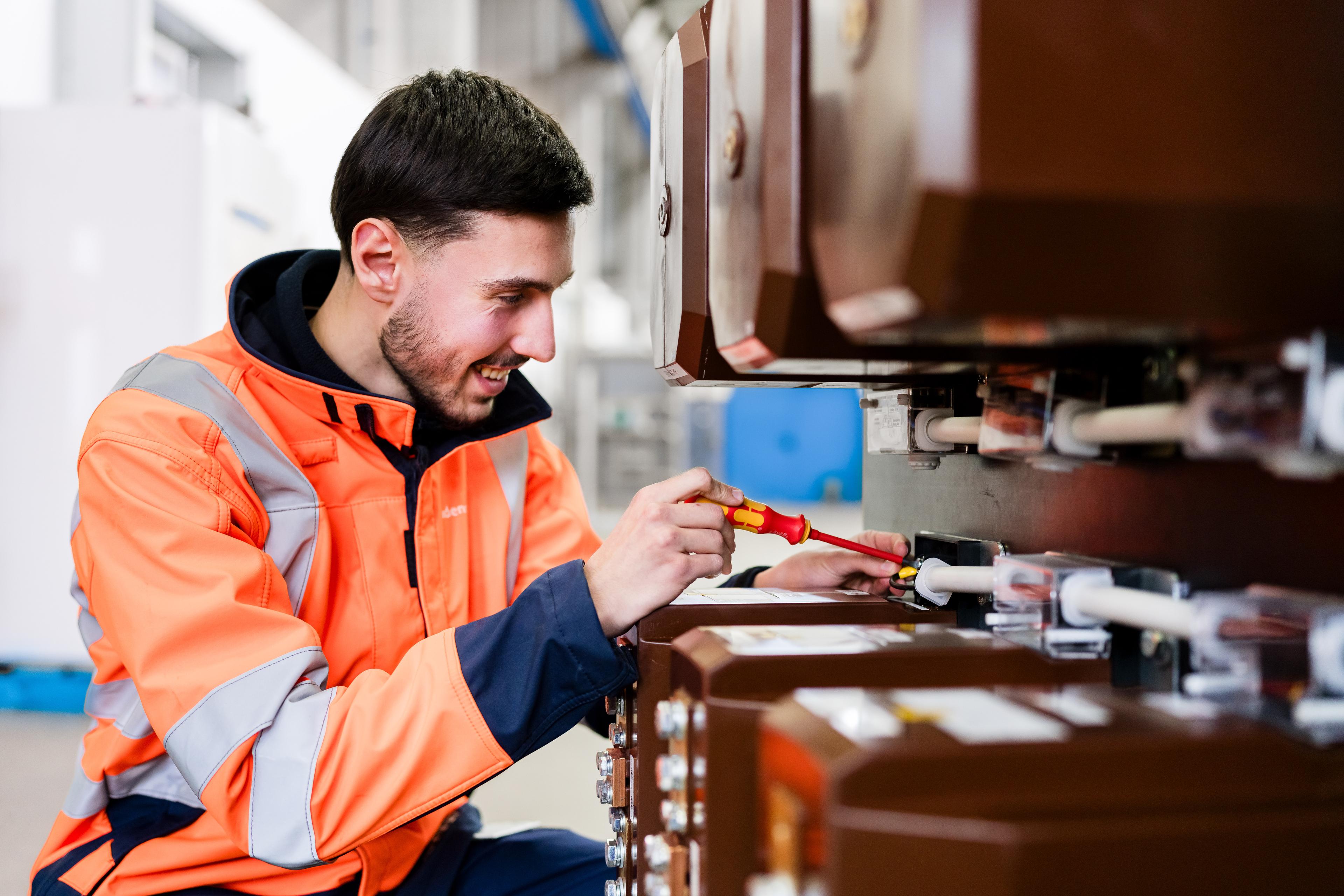 Fachkraft im Badenova kundenservice in orangefarbener Warnschutzjacke arbeitet mit Schraubendreher an einer industriellen Anlage. Nahaufnahme zeigt präzise Handarbeit an technischen Komponenten in einer Produktions- oder Wartungsumgebung.