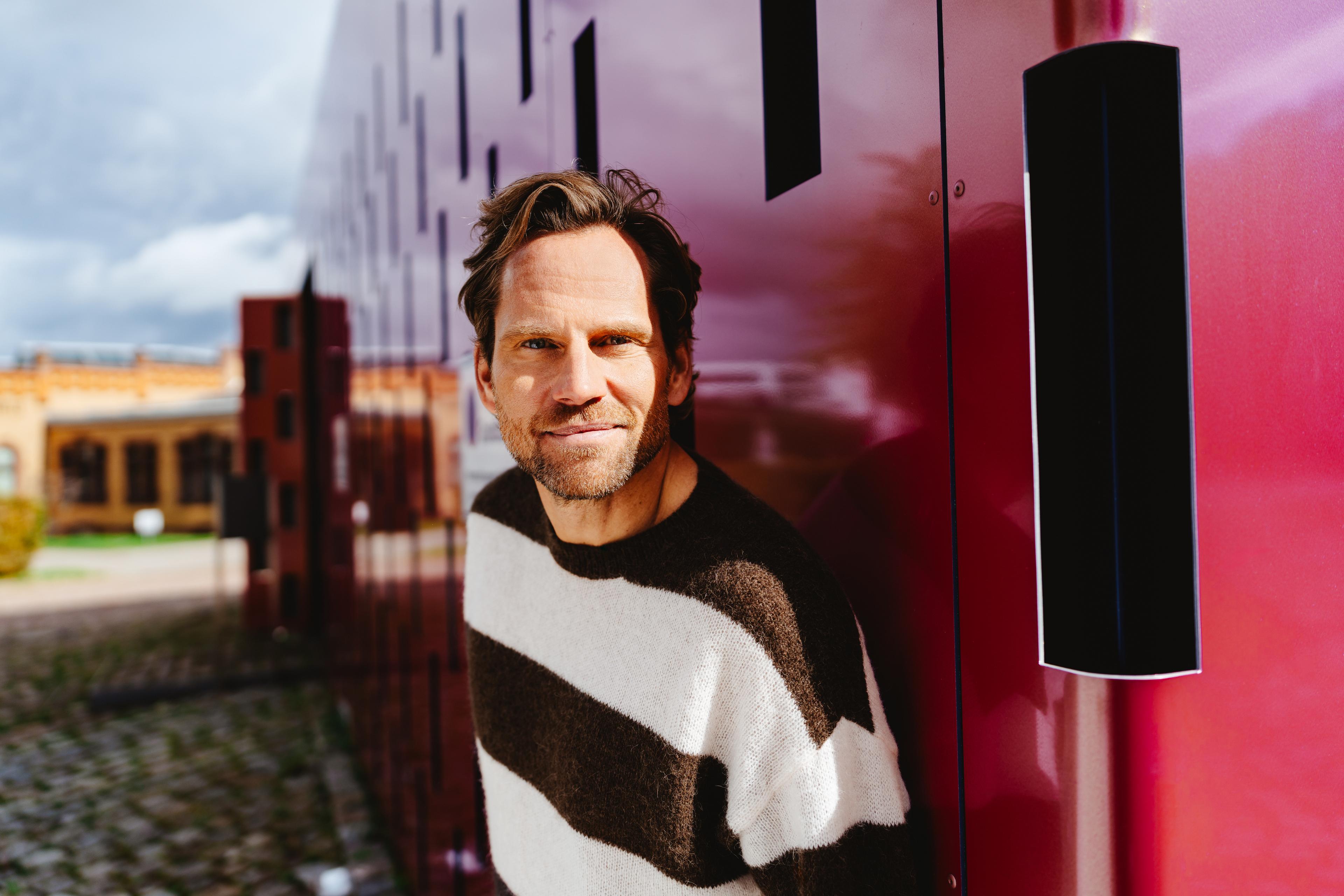 Mann mittleren Alters steht vor einem roten Gebäude und schaut in die Kamera