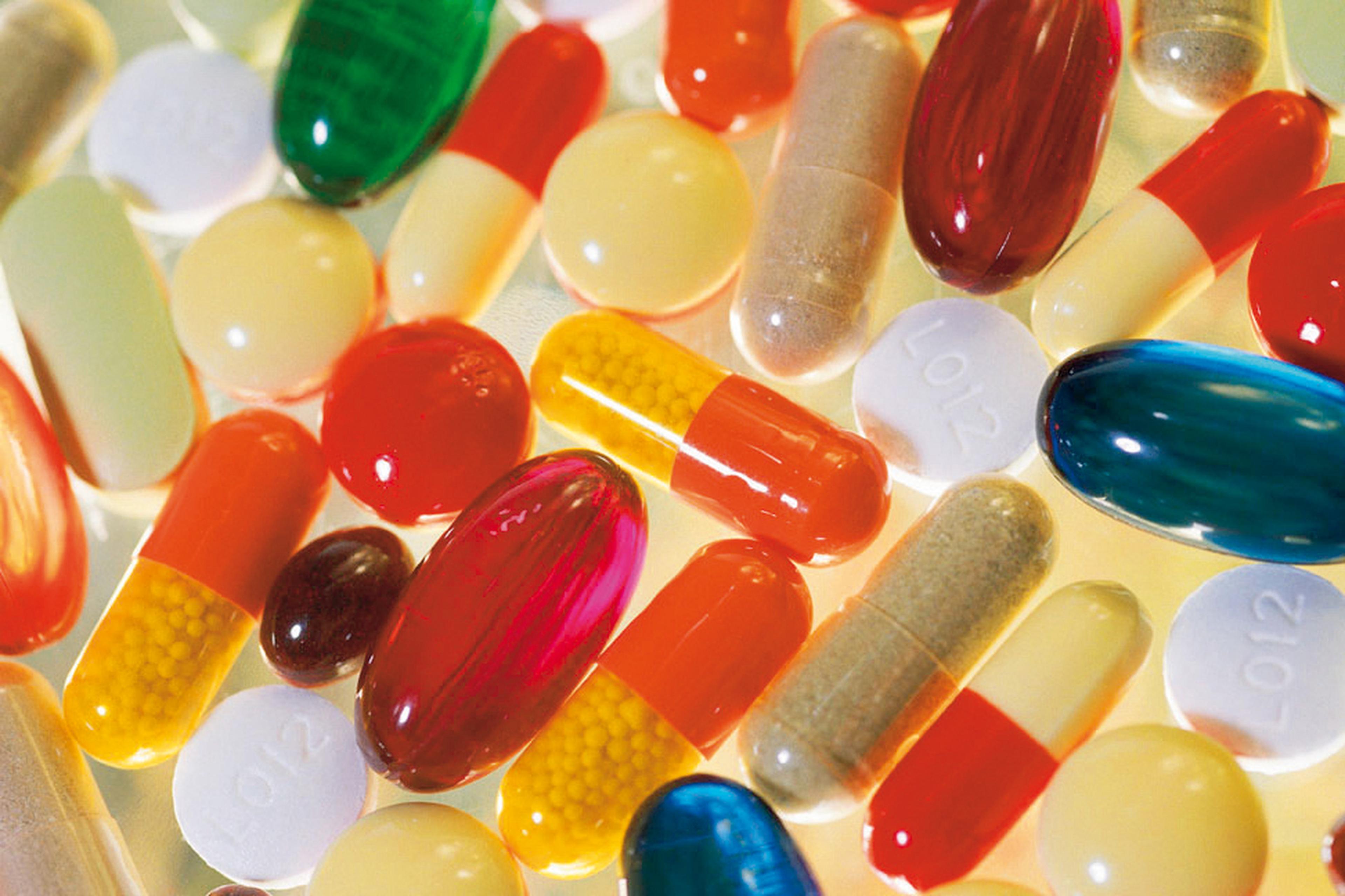 Auf einer spiegelnden Oberfläche liegen verschiedene bunte Tabletten und Kapseln verstreut, darunter rote, blaue, gelbe und weiße.