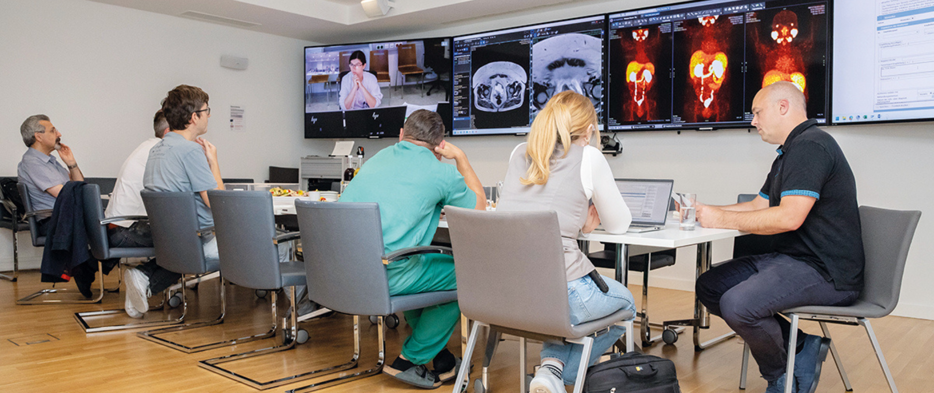 People in a meeting room reviewing medical scans on large screens. Some are seated at a table with laptops, while others observe.