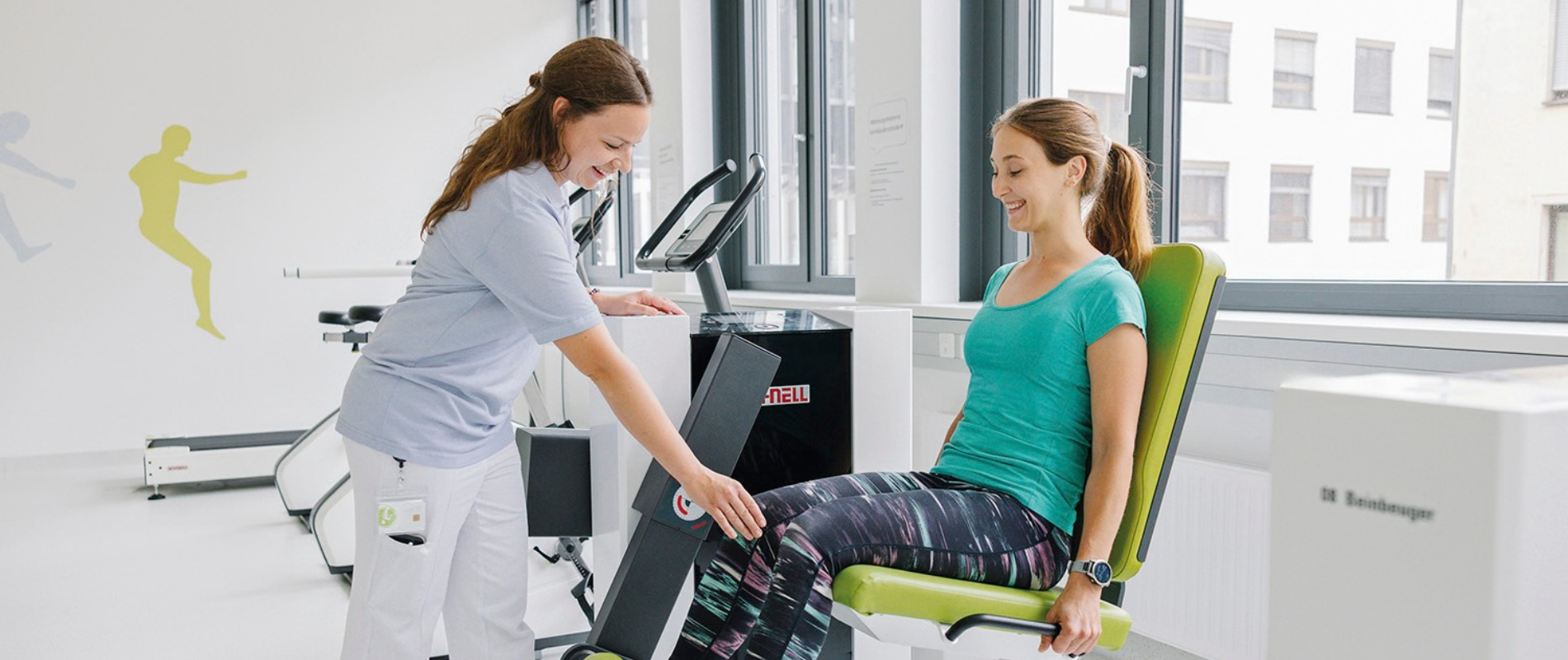 Therapeutin erklärt Patientin auf Trainingsgerät Übung.