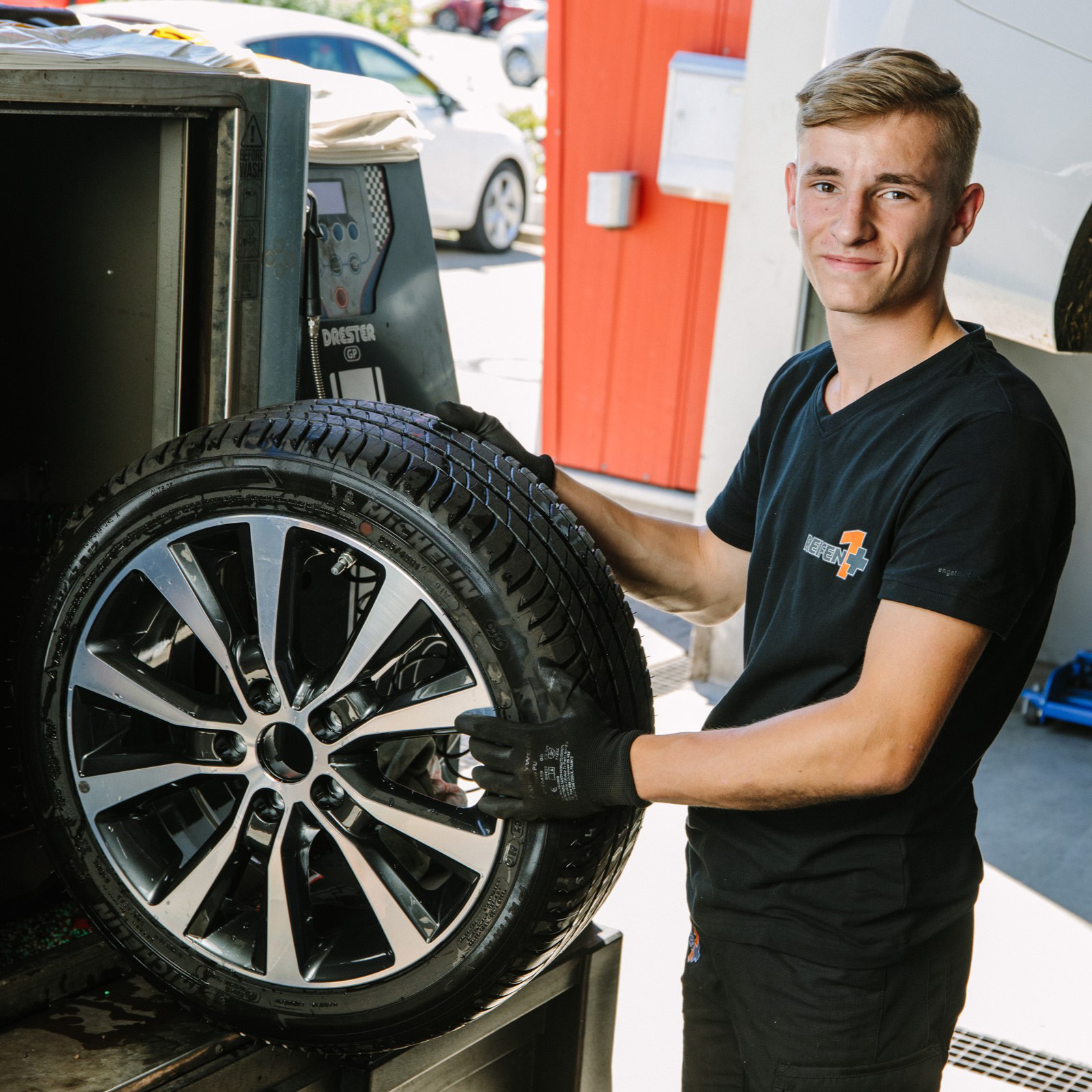 A person in a workshop holding a car tire with a shiny alloy wheel, wearing gloves and a black shirt, standing next to equipment.