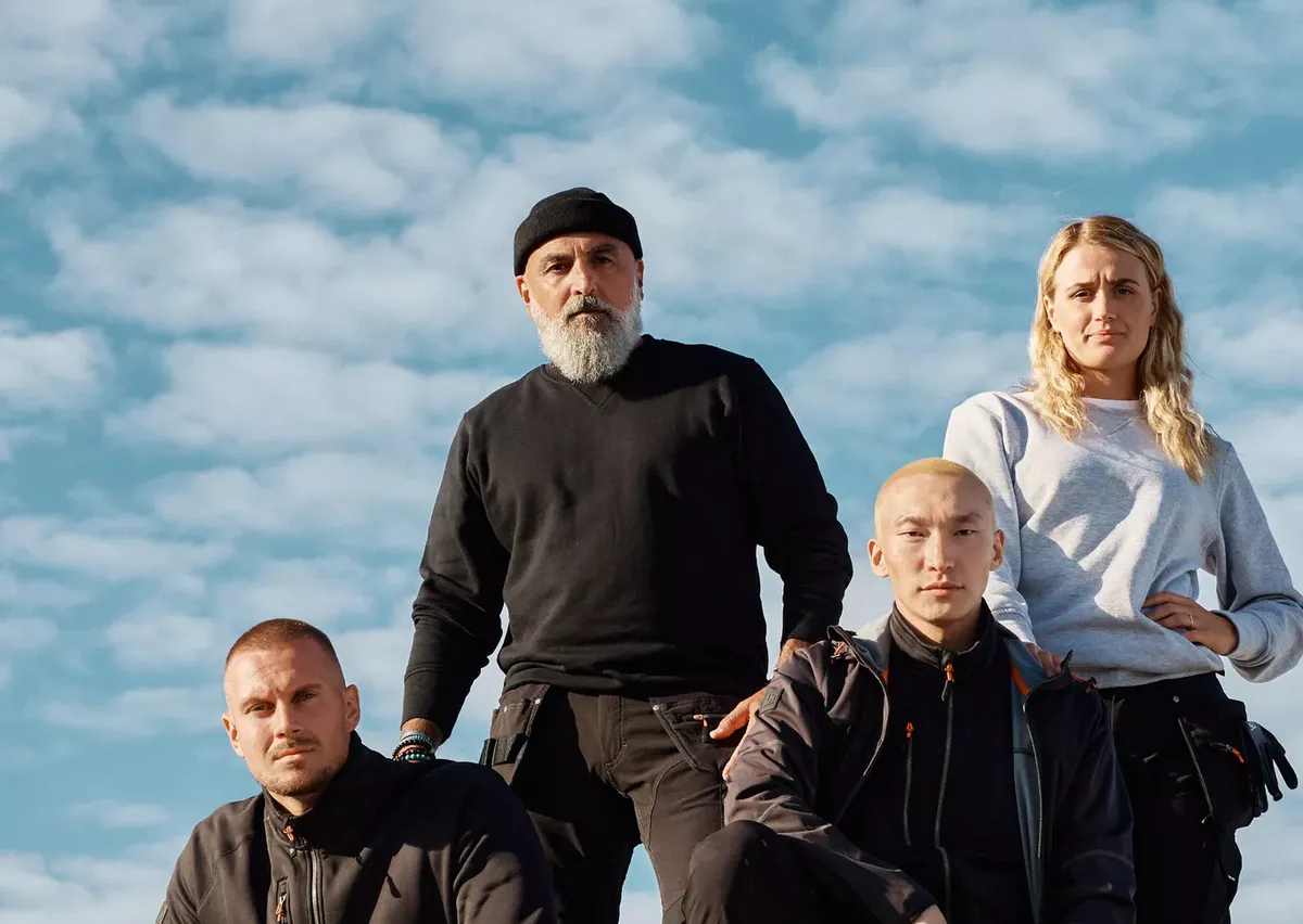 Four people in casual attire pose confidently on a concrete ledge against a partly cloudy sky.