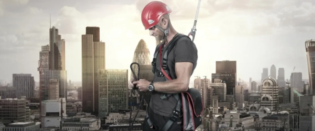 En byggarbetare med röd hjälm och skyddsutrustning står på ett tak med stadens silhuett i bakgrunden.