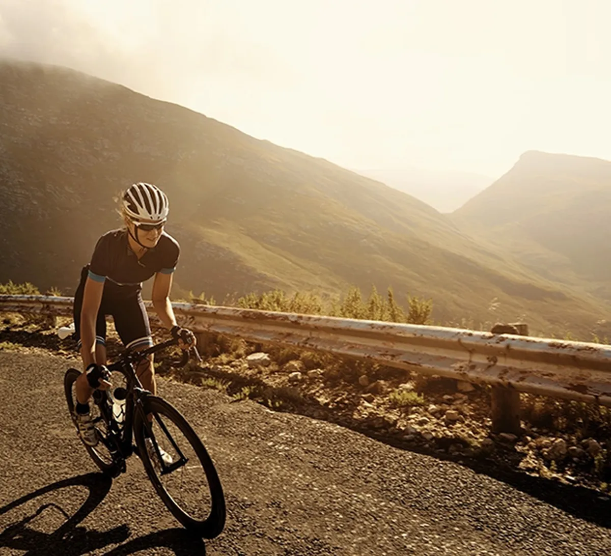 Cyclist in a helmet rides uphill on a mountain road during sunset, with a scenic view of hills and a metal barrier in the background.