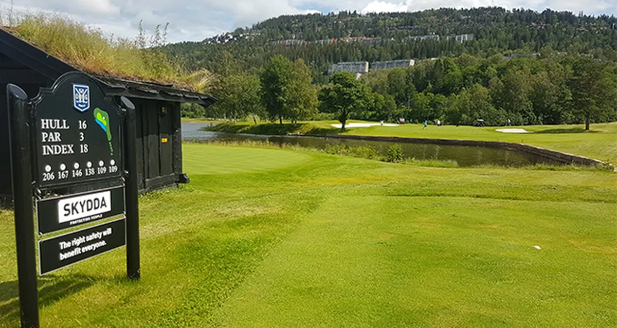 Par‑3 tee at hole 16 on a lush golf course with wooden sign and turf‑roof shelter, short pond, green and tree‑covered hillside beyond.