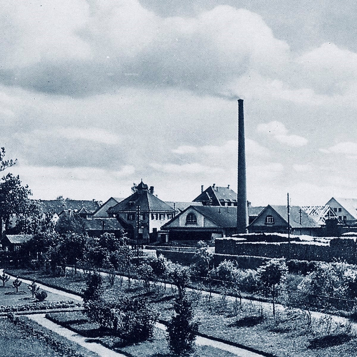A black and white image of a factory with a tall chimney, surrounded by trees and a garden.